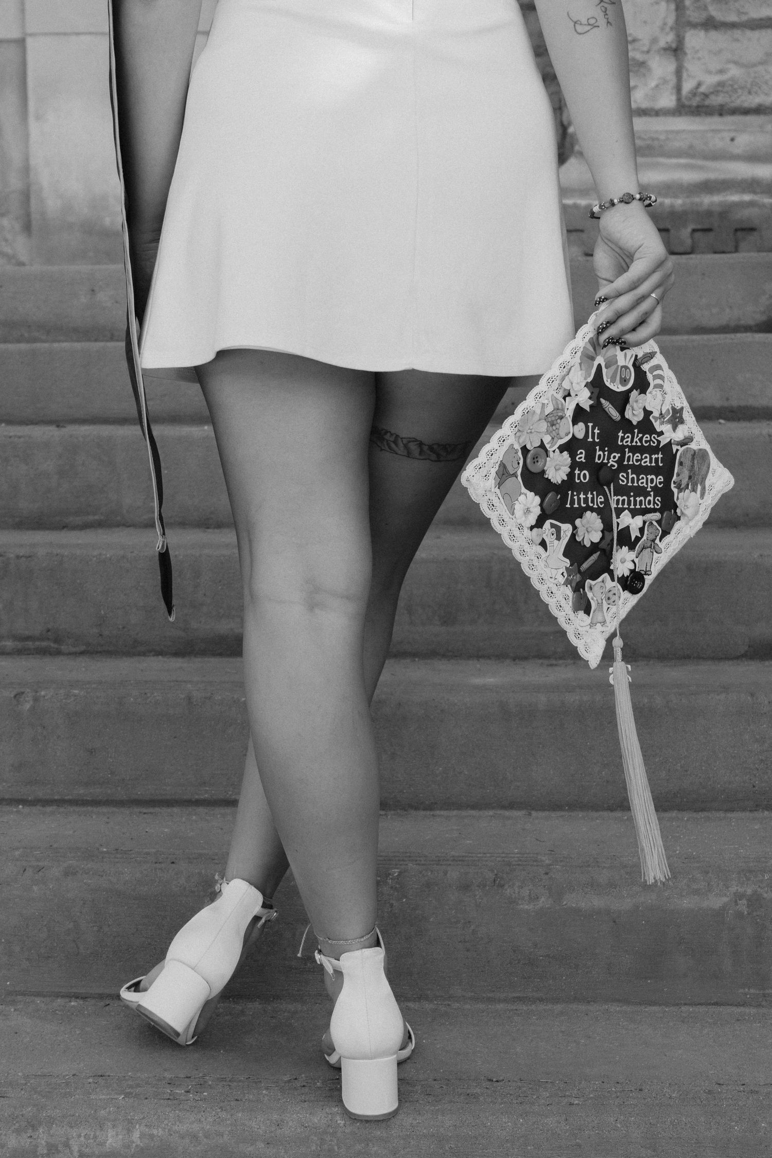 Black and white photo of a graduate holding decorated cap on steps, wearing white dress and heeled sandals.