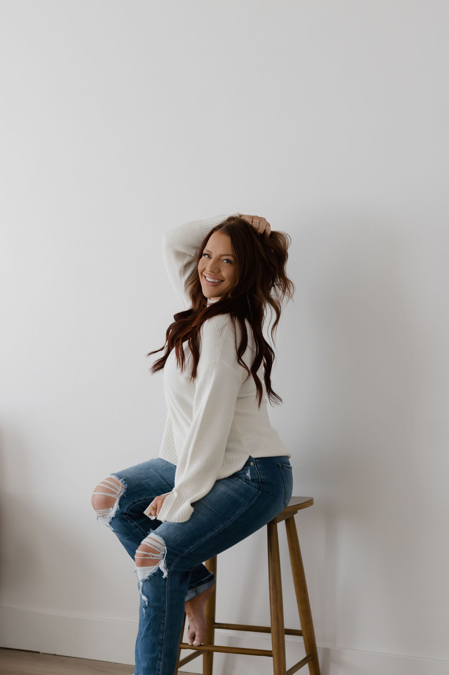 A person in a white sweater and ripped jeans poses casually on a wooden stool against a white wall.
