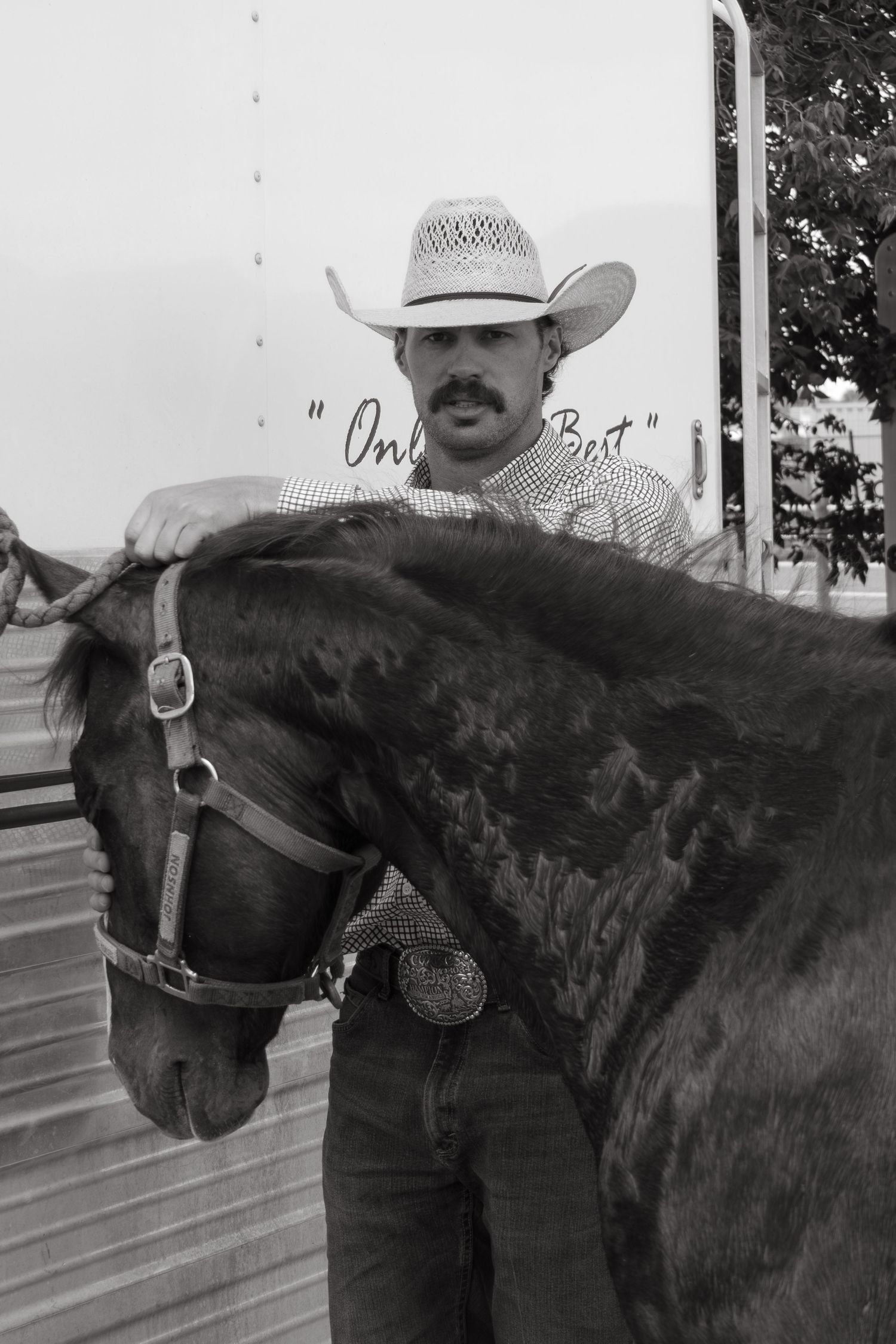 Black and white photo of a cowboy with mustache wearing a straw hat standing beside a dark horse with bridle.