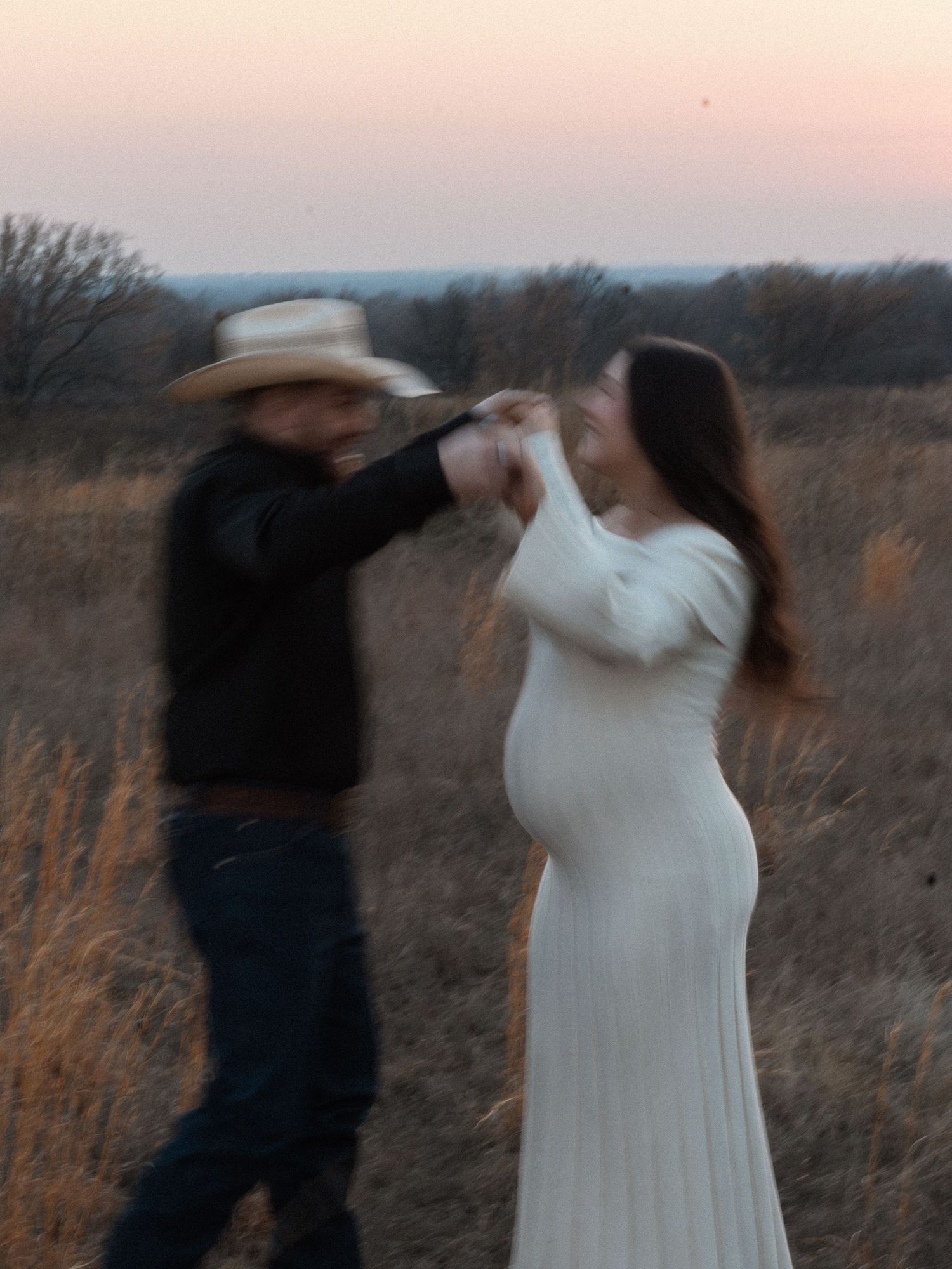 Couple dancing in a field at dusk, pregnant woman in white dress with man in cowboy hat, motion blur effect.