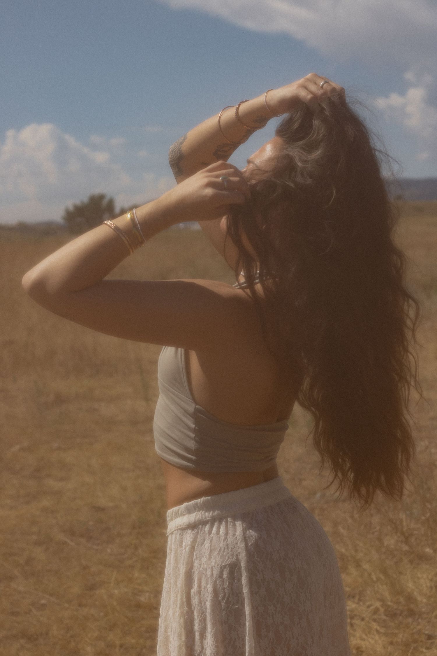 Woman with long curly hair poses in a golden field, wearing a gray crop top and white skirt under a blue sky.