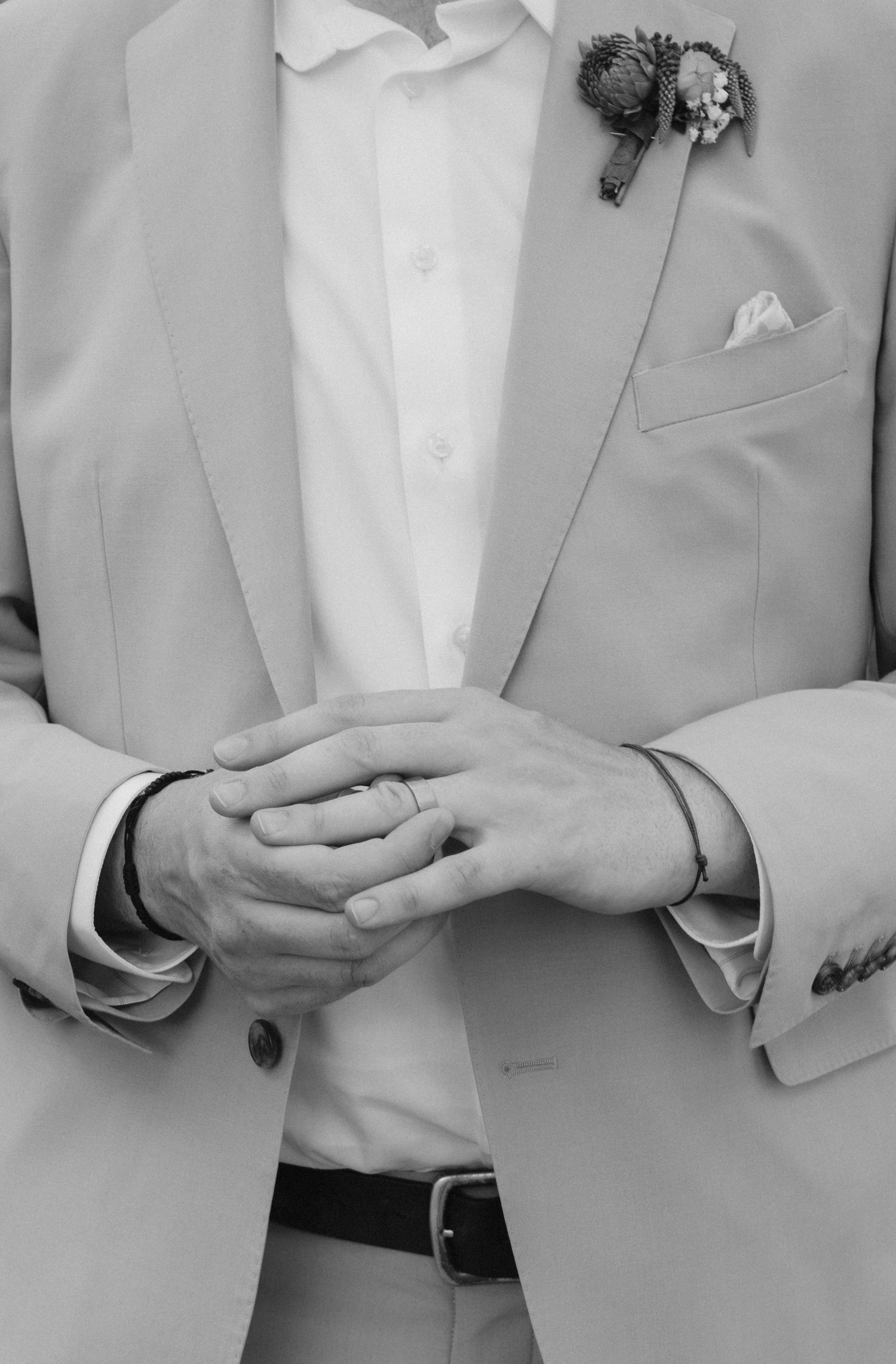 Black and white photo of a groom in a light suit with boutonniere, clasping hands showing wedding ring.