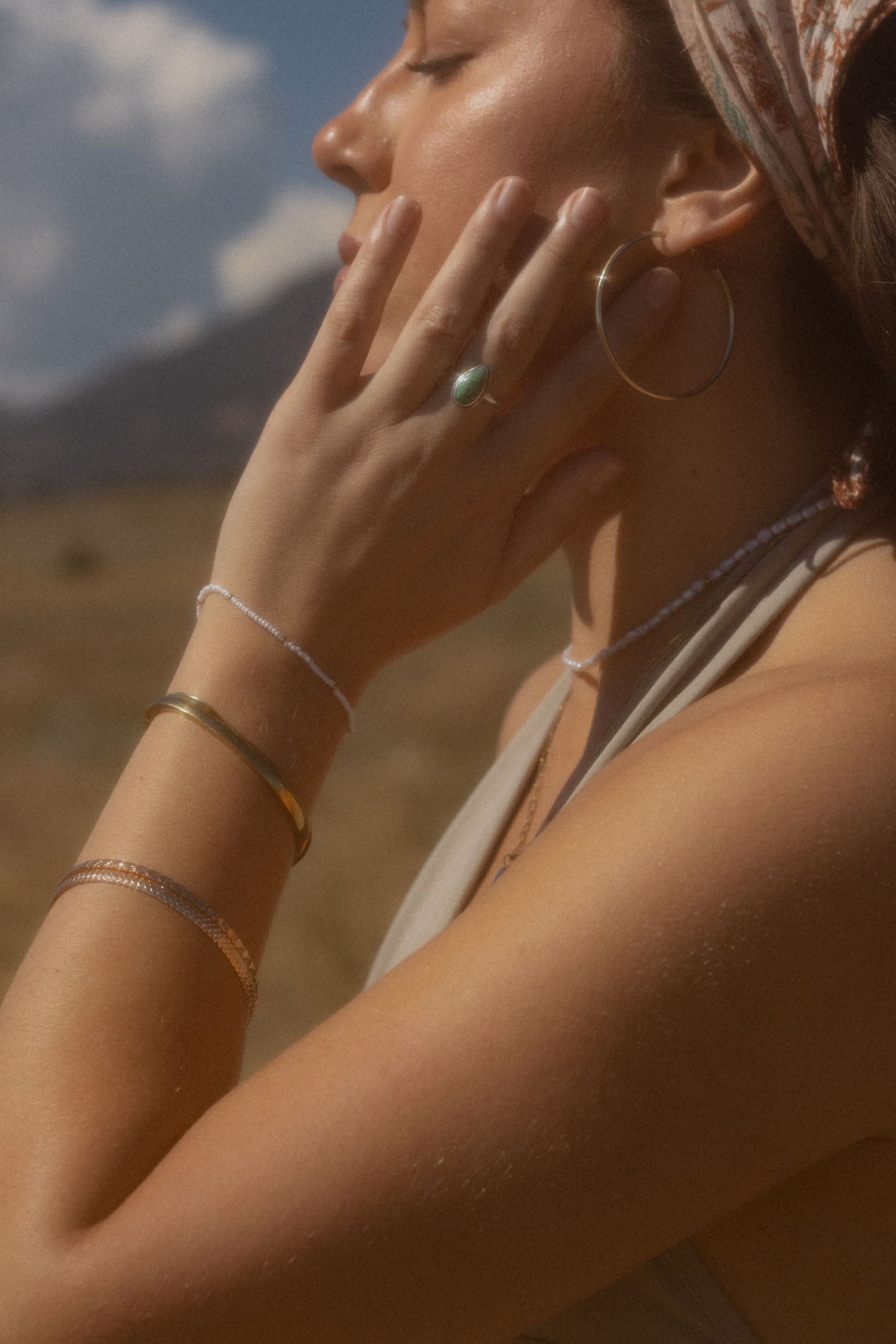 Woman wearing gold hoop earrings, turquoise ring, and layered bracelets outdoors with mountains in background.