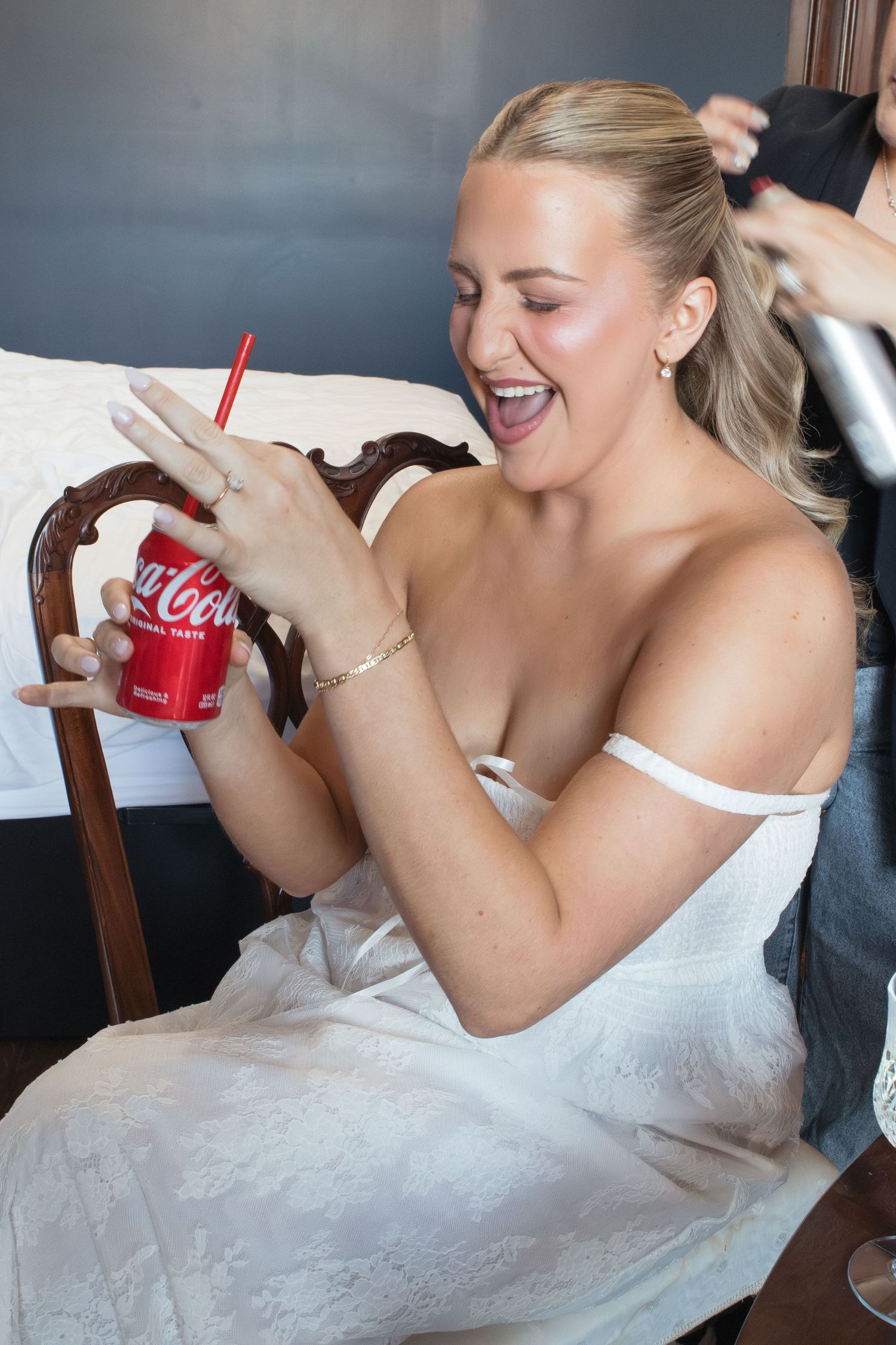 Bride in white dress holding Coca-Cola can laughs while getting hair styled before wedding ceremony.