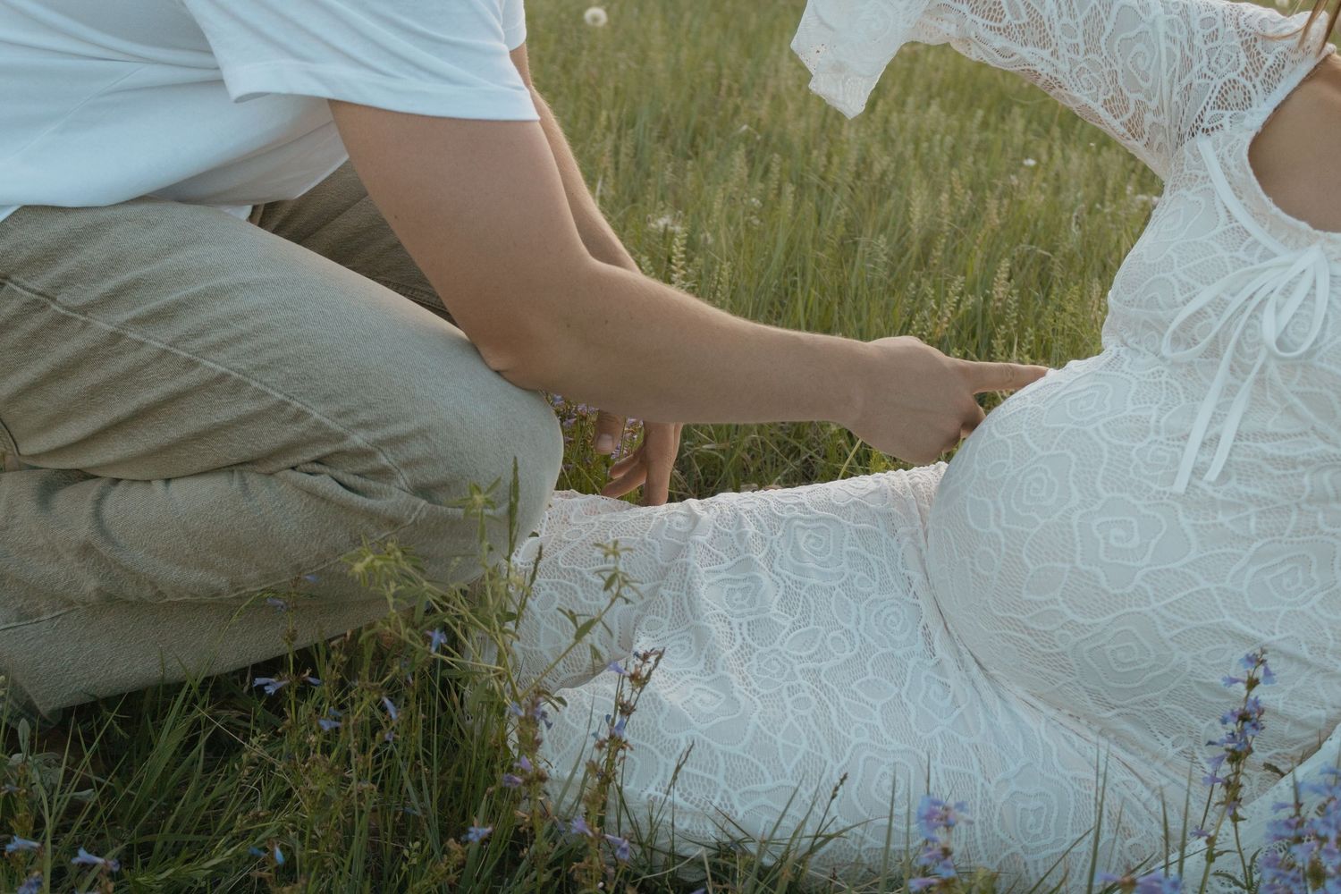 A couple sits in a lush green meadow, tenderly holding hands, with the woman in a white dress.