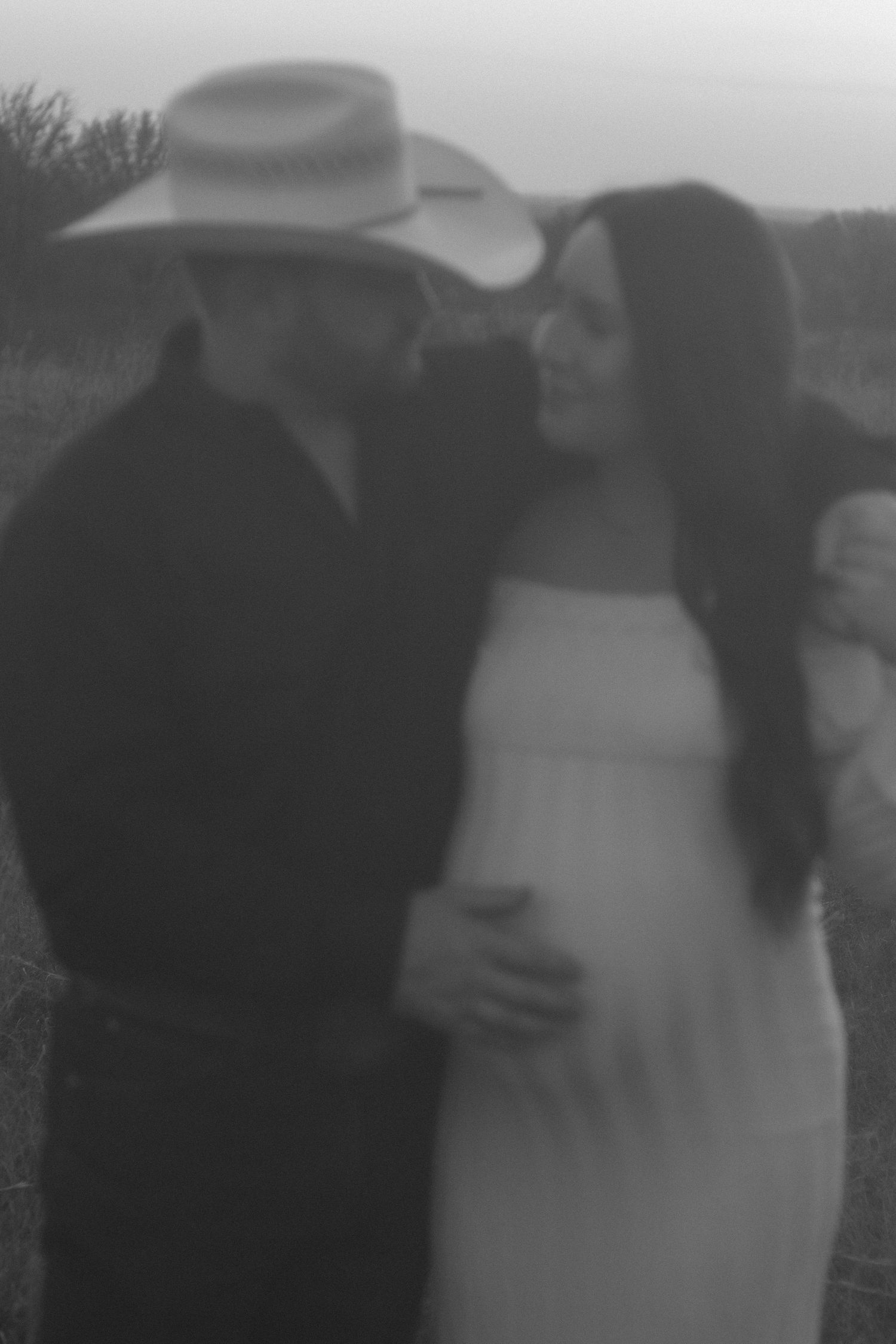 Black and white photo of a couple in a field, man in cowboy hat embracing pregnant woman in white dress.