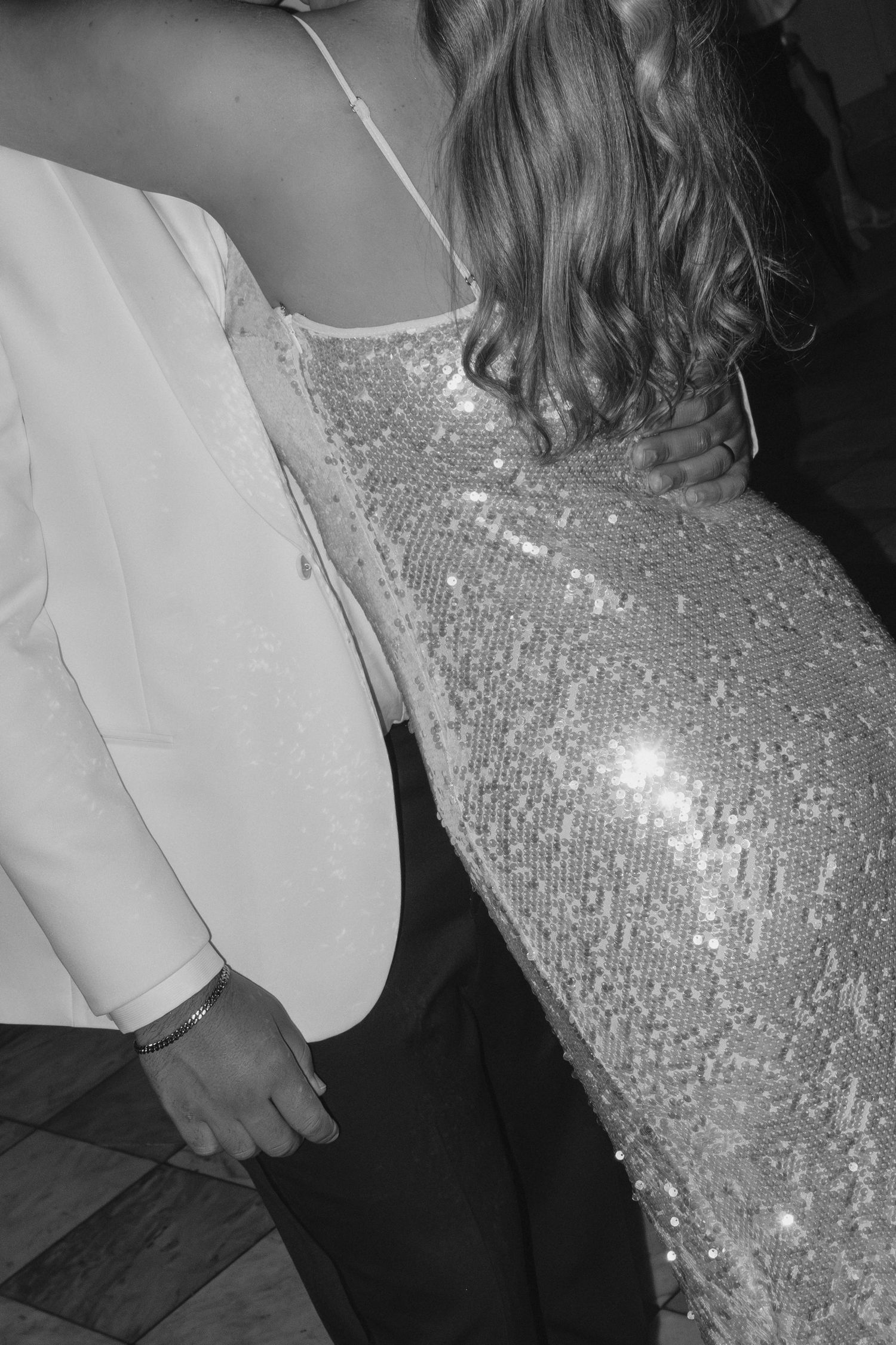 Black and white photo of couple dancing, woman in sequined dress and man in white blazer holding hands.