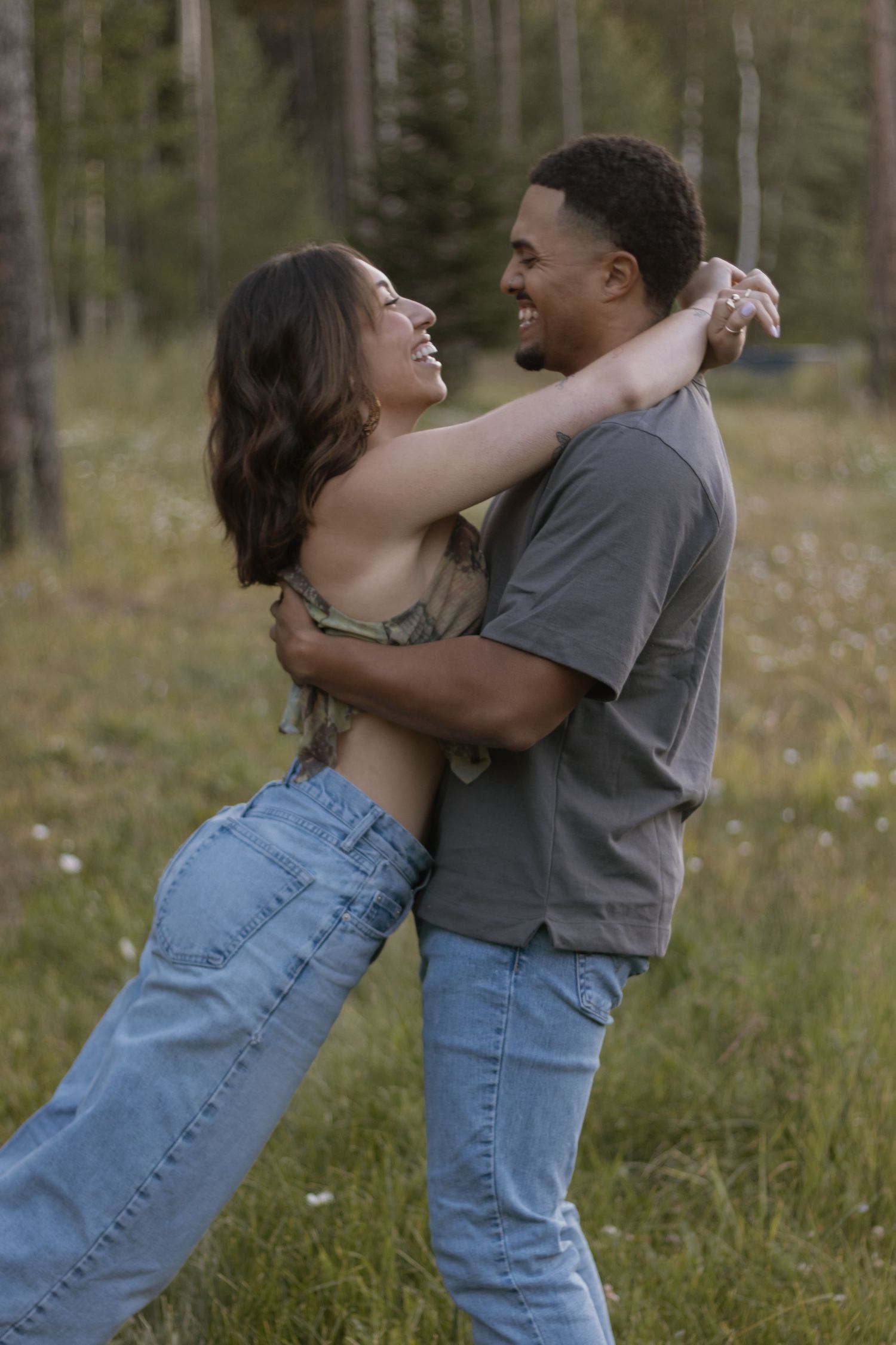 A couple embraces playfully in a grassy field wearing casual denim and gray clothing.