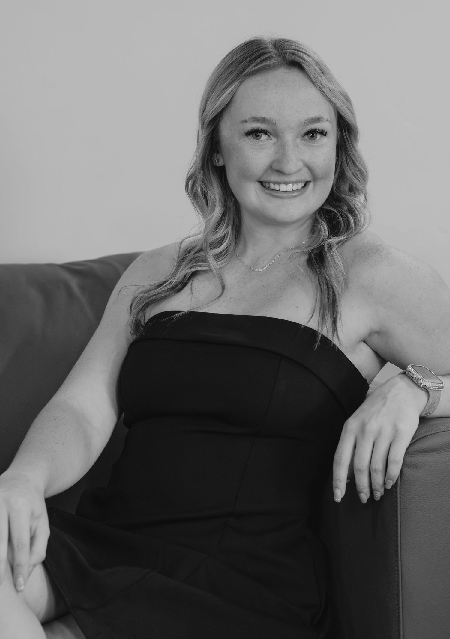 Smiling blonde woman in black strapless dress sitting relaxed on sofa, black and white portrait photo.
