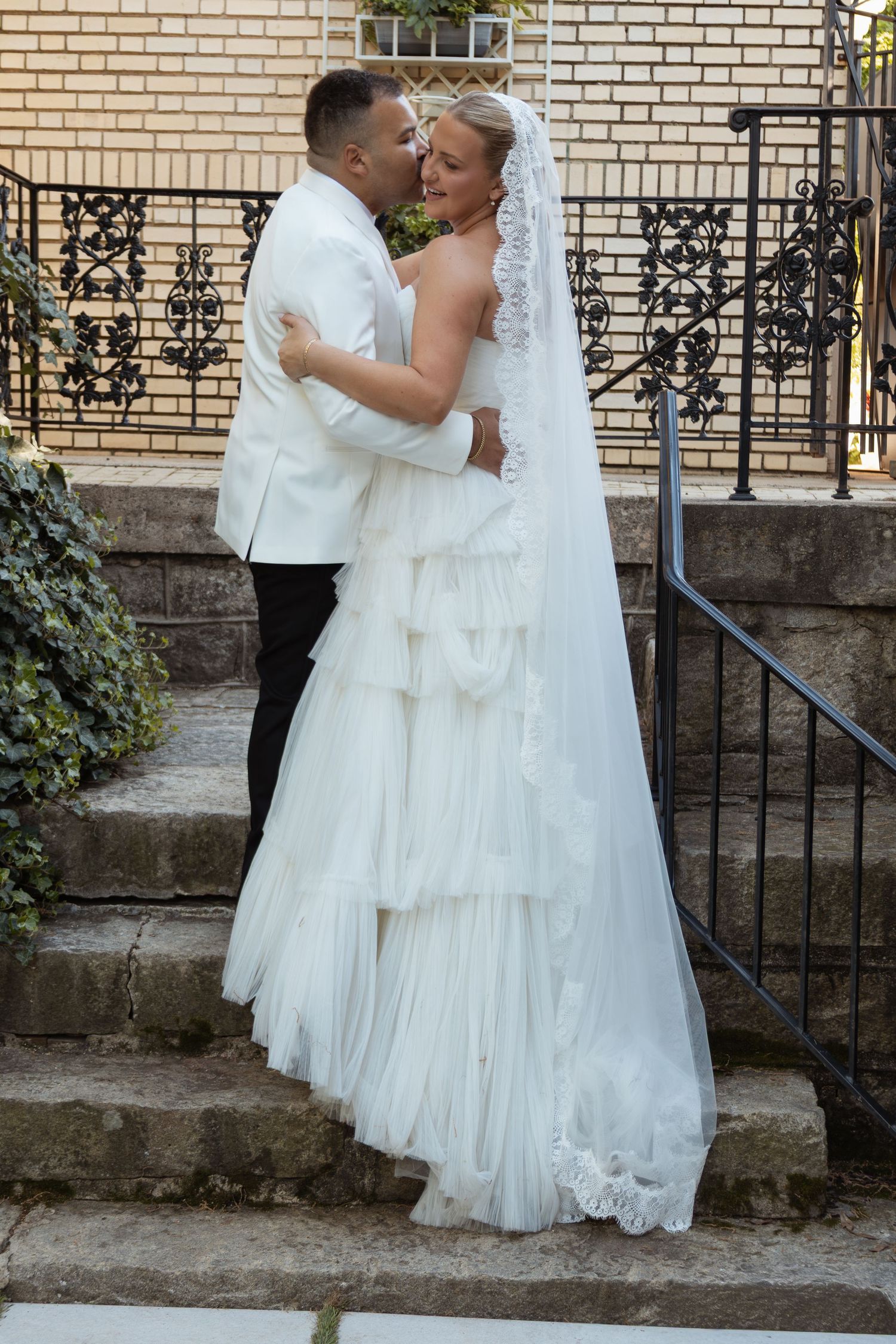 Bride in white ruffled gown and veil shares a romantic kiss with groom in white suit on stone steps.