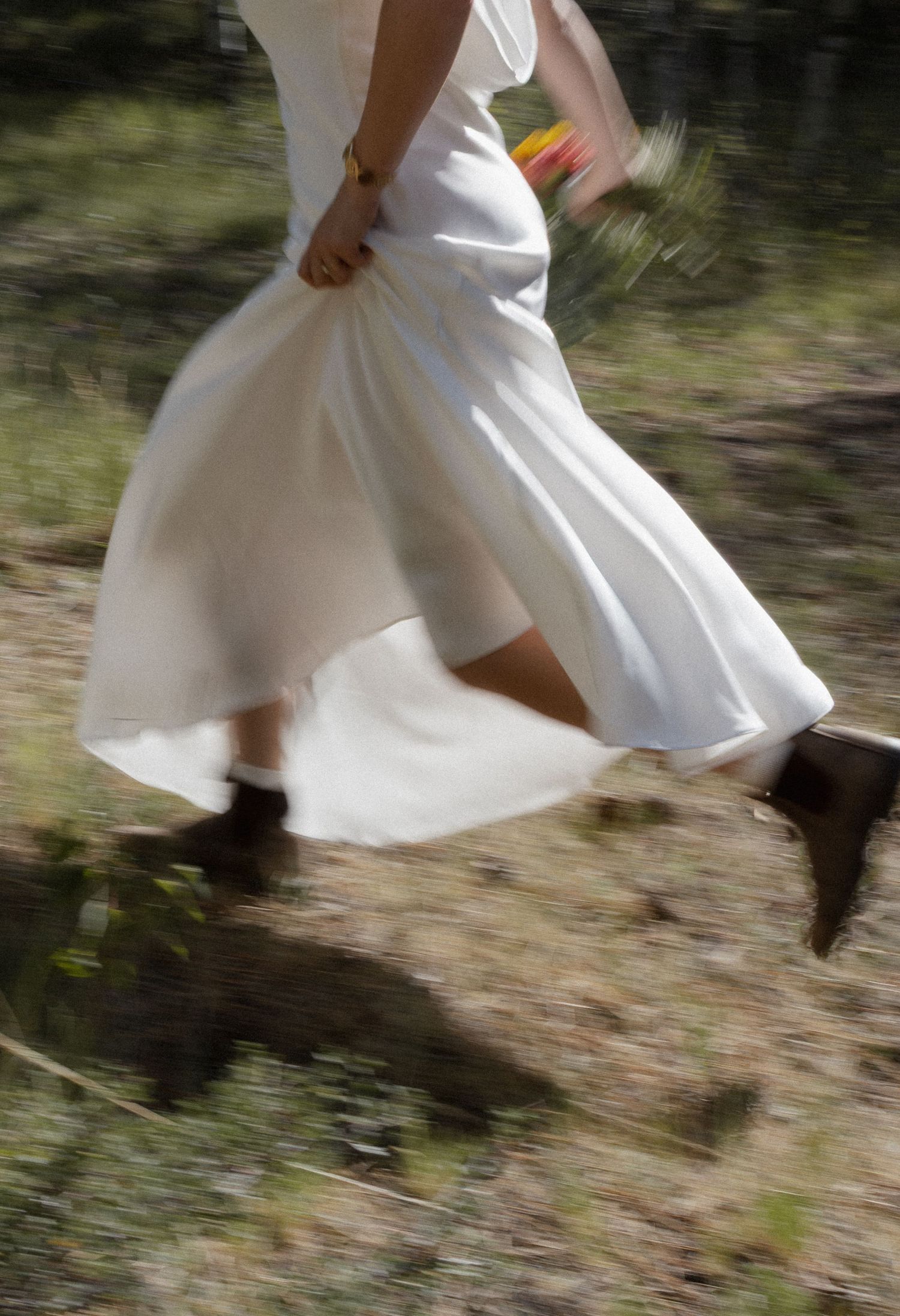 Bride in white wedding dress and brown boots running through a natural outdoor setting, holding a colorful bouquet.