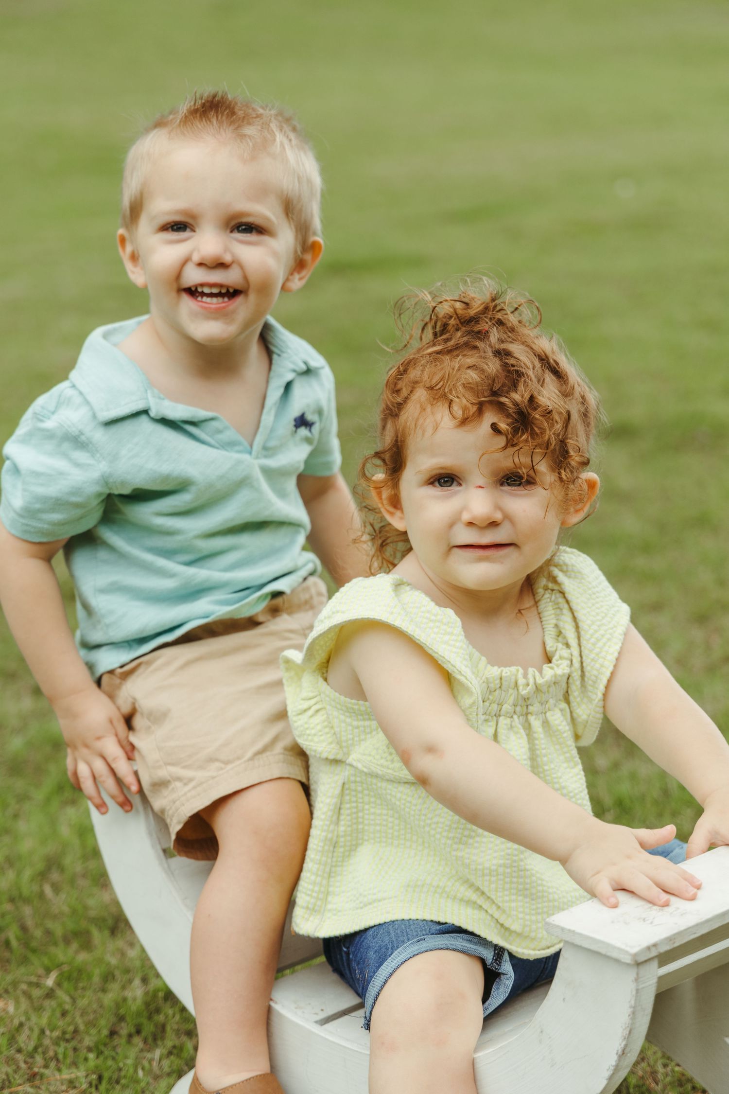 Two small children sit together on a white chair outdoors.