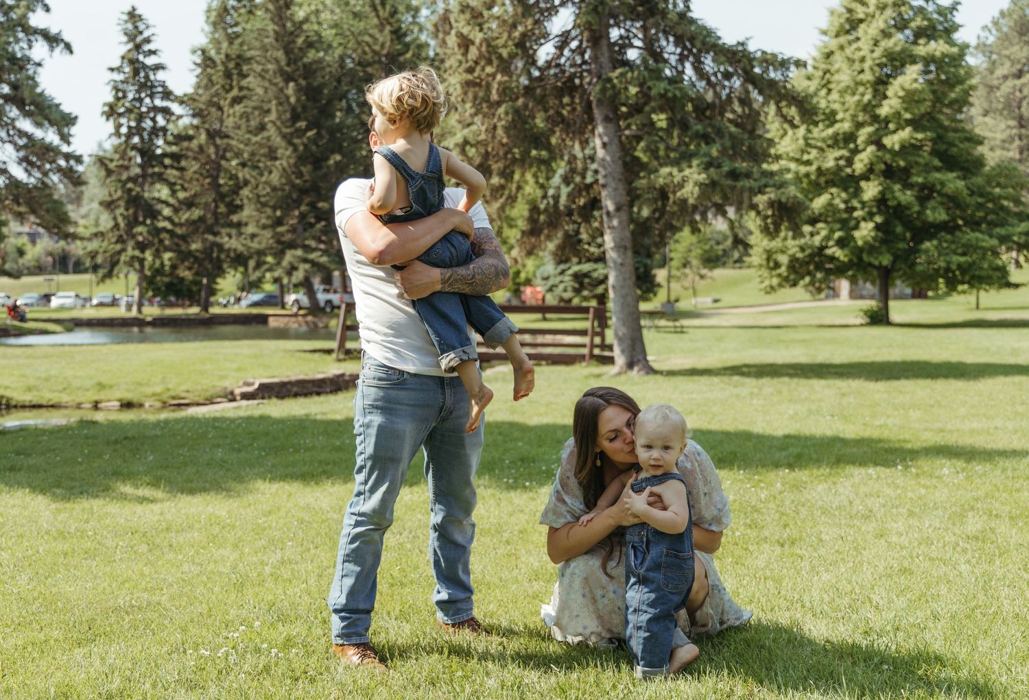 A family walks together through a green park with tall pine trees in the background.