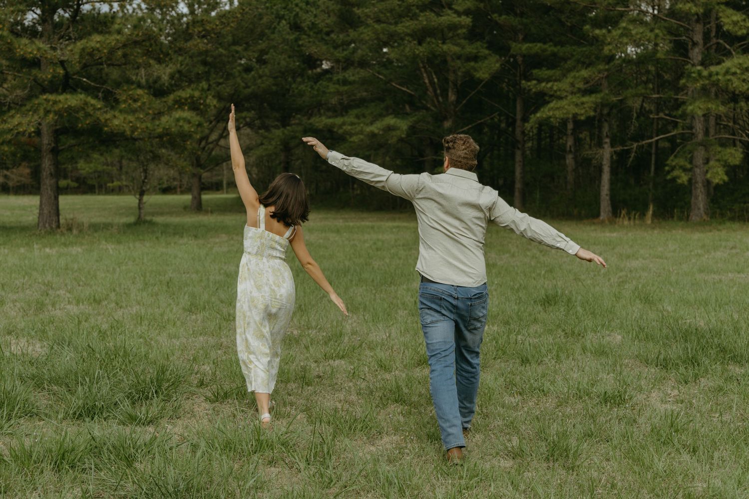 A couple dances playfully in a grassy field with trees in the background.
