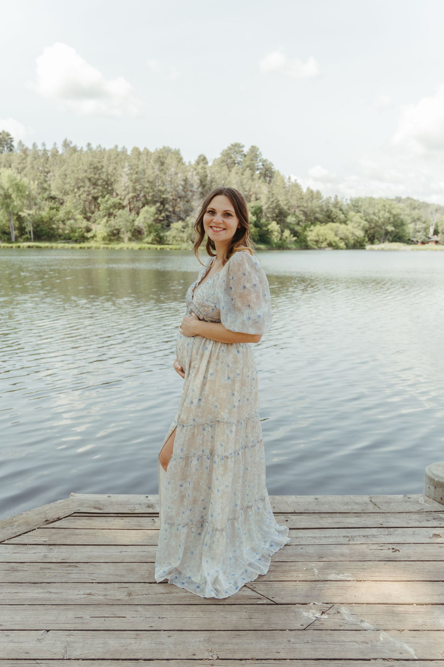 A woman in a flowing lace dress poses gracefully on a wooden dock by a serene lake.