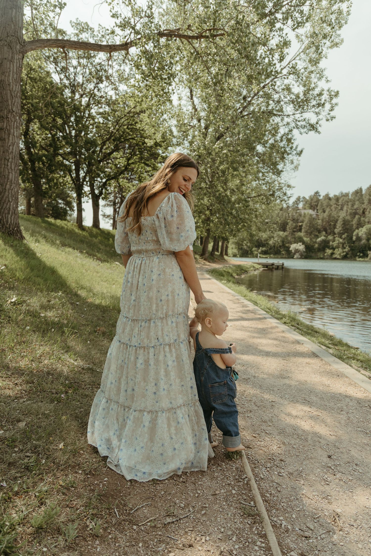 A family walks along a lakeside path surrounded by trees on a sunny day.