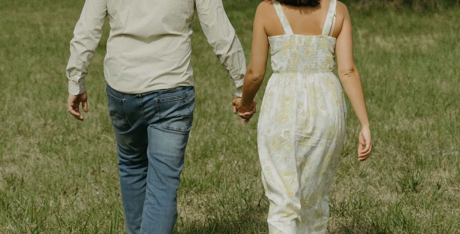 A couple walks hand in hand through a sunny meadow.