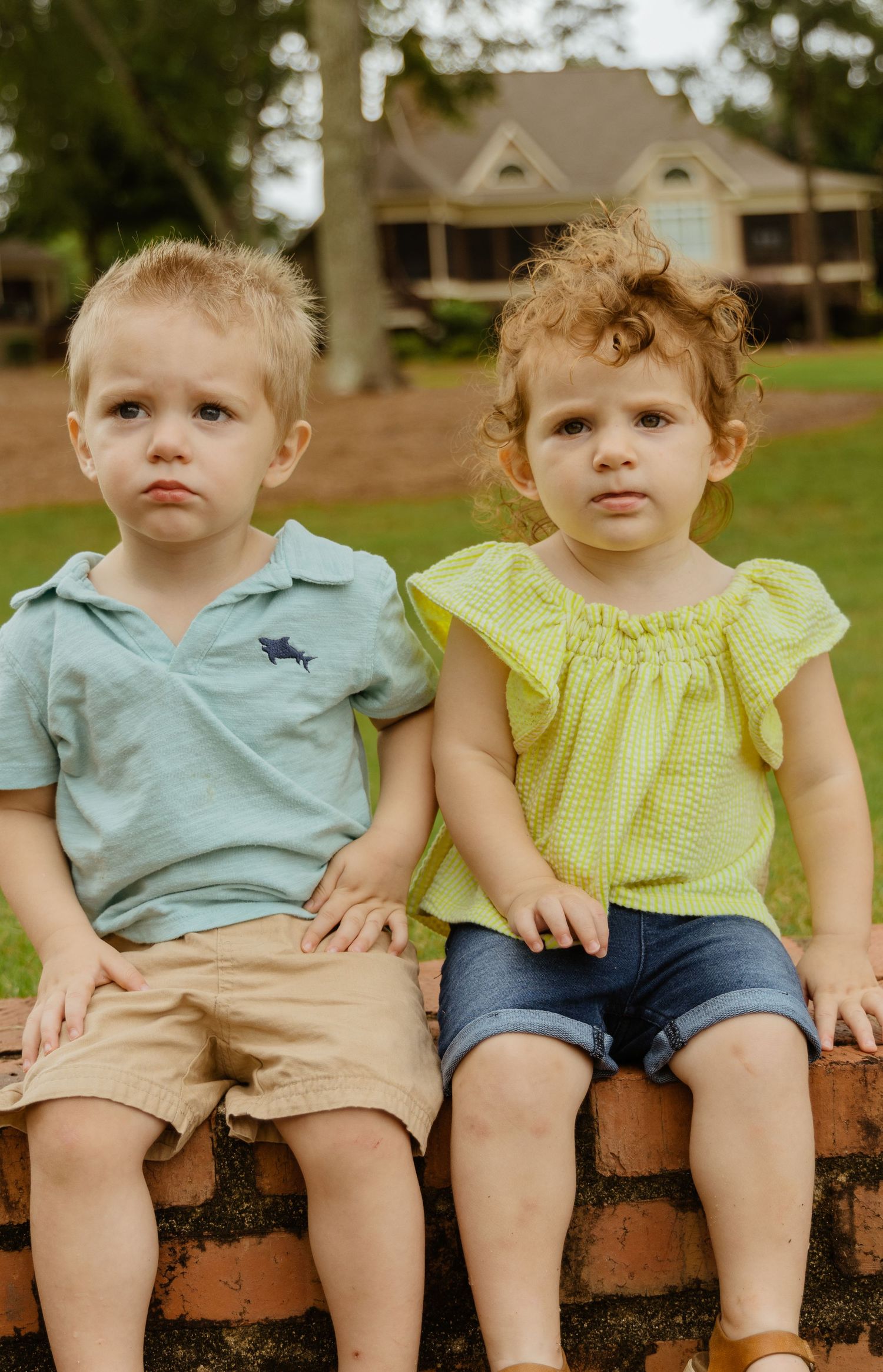 Two children sitting together on brick wall wearing summer outfits.