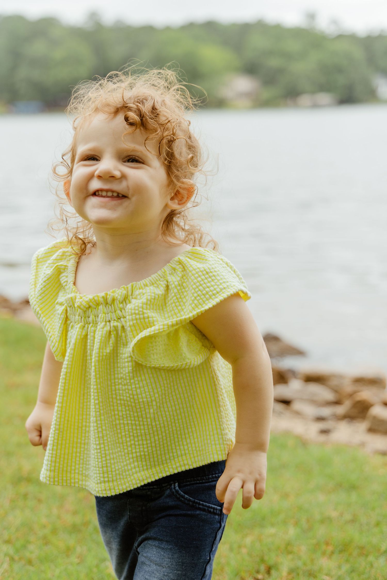 Child in yellow top smiling and playing outdoors near water.