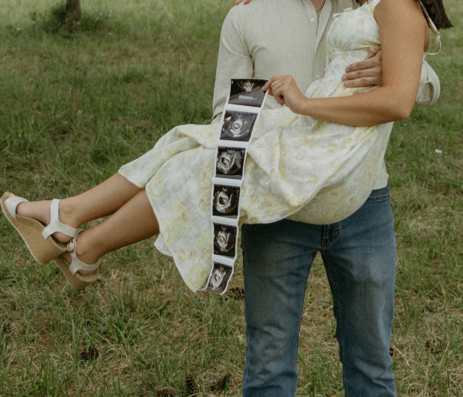 A couple holds ultrasound photos while standing in a grassy field.
