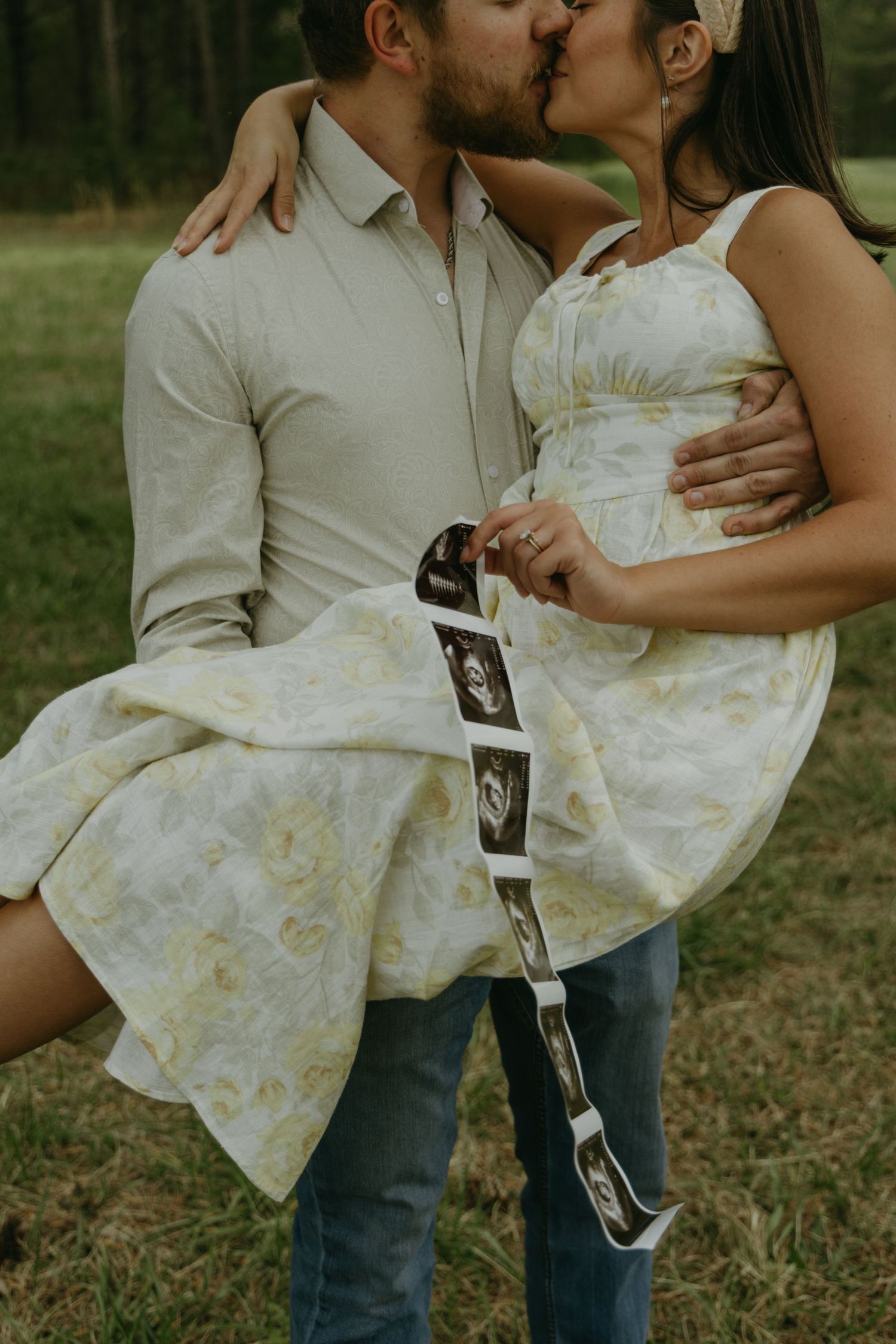 A couple holds ultrasound photos while embracing in a grassy field.