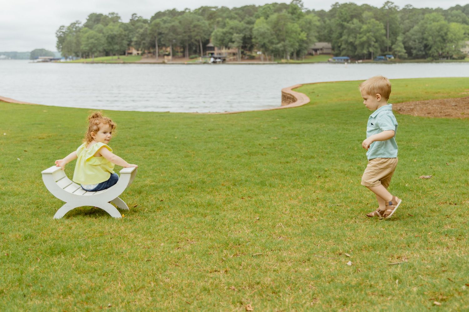 Children running and playing on grass near a lake on a sunny day.