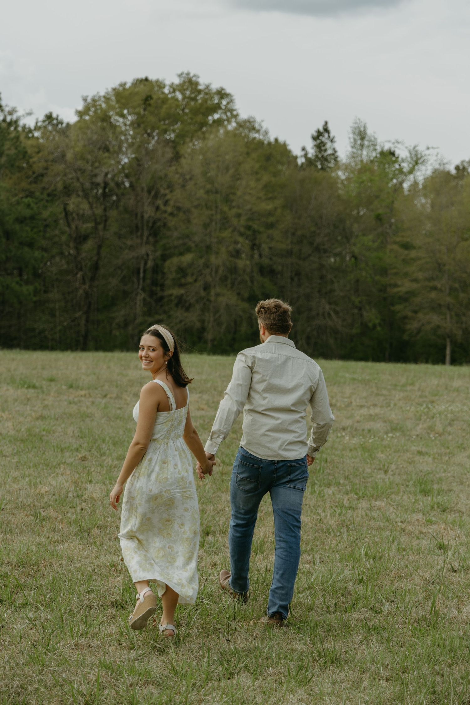 A couple walks away through a grassy field on a cloudy day.