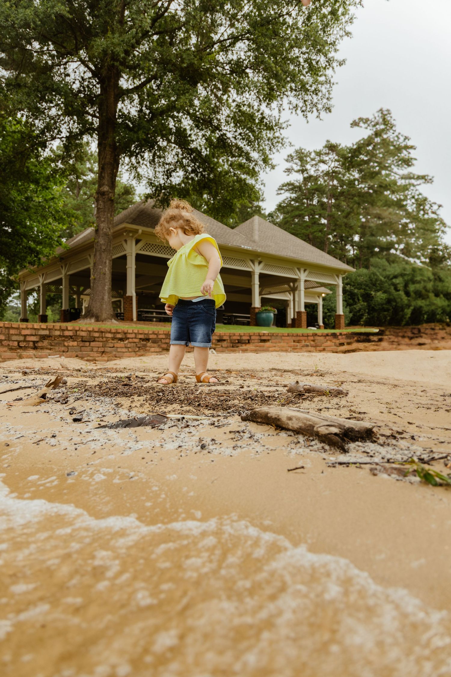 Child in yellow top plays near muddy shoreline with house in background.