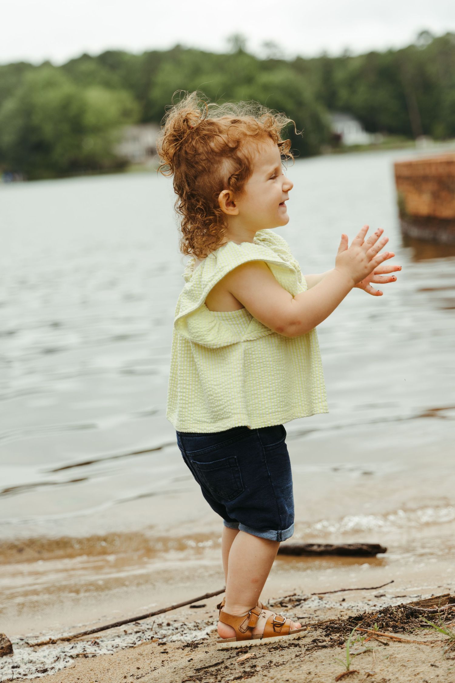 Child in yellow ruffled top stands by water's edge clapping hands.