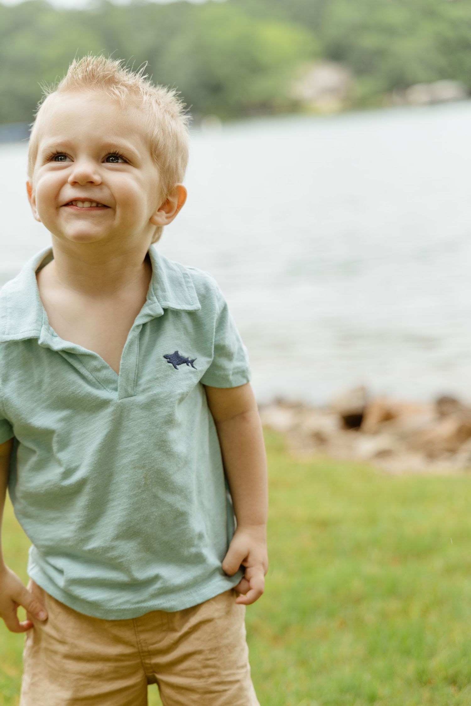 A young child in a mint green polo shirt smiles joyfully outdoors near a lake.