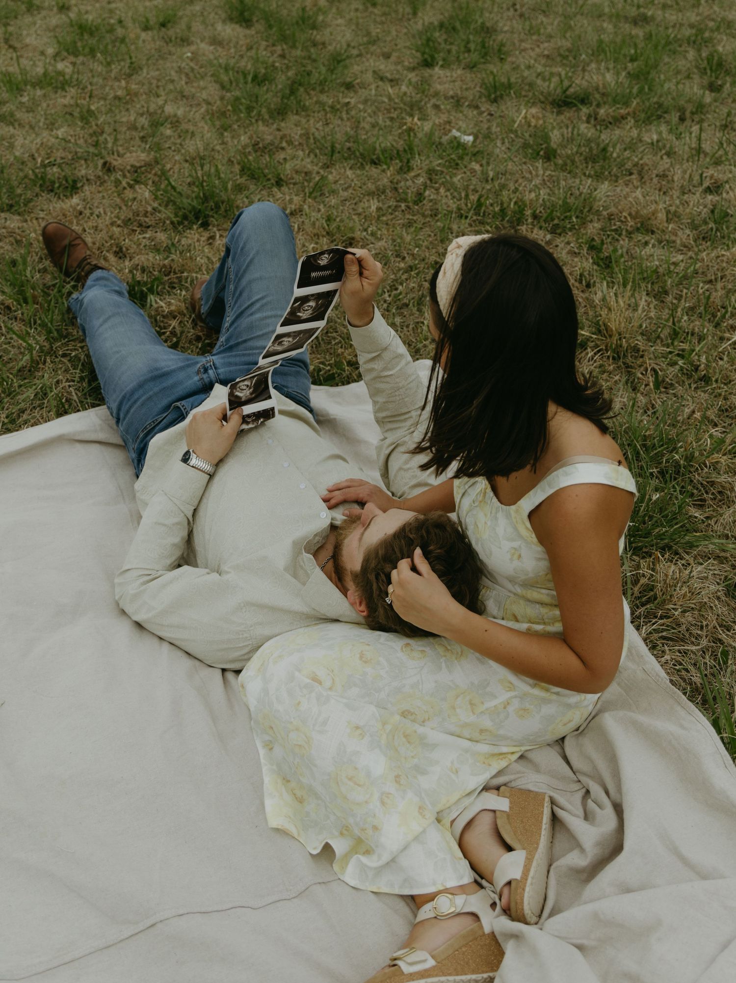 A couple lies on a white blanket in a grassy field during their pregnancy photoshoot.