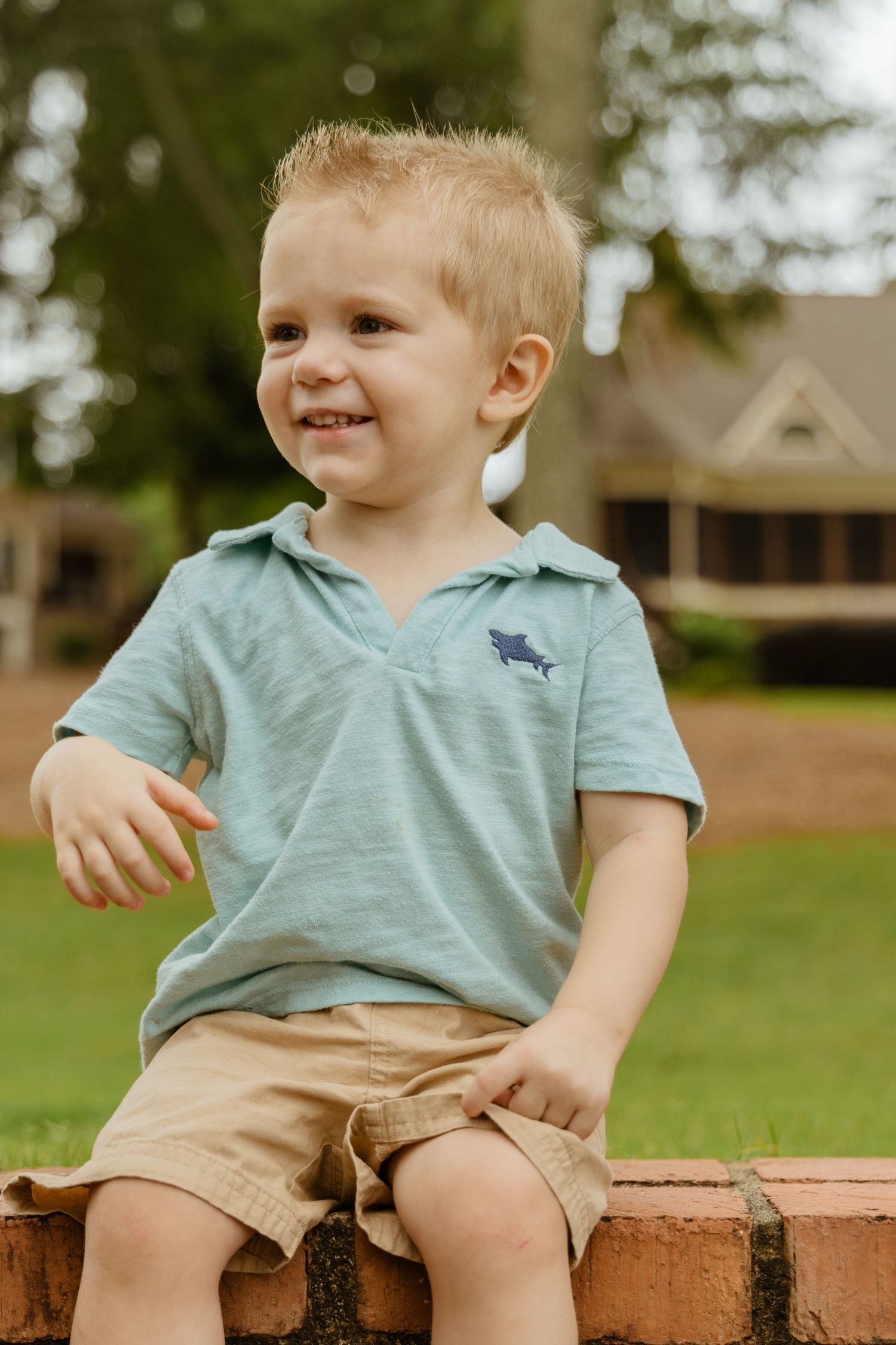 Child in mint polo shirt sitting on brick wall with house in background.