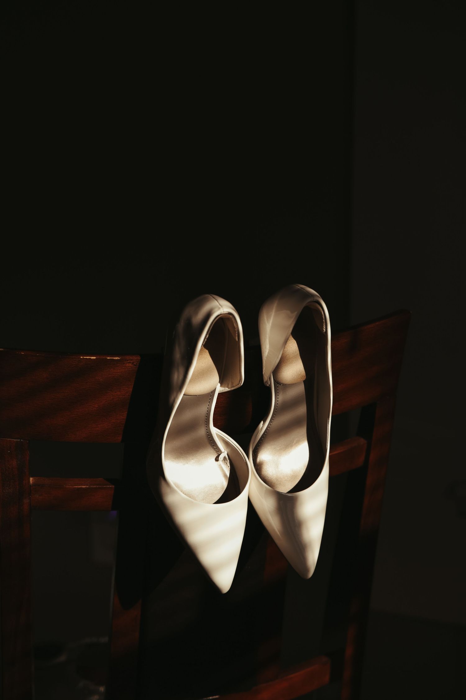 Elegant white bridal heels photographed in dramatic lighting on a wooden surface.