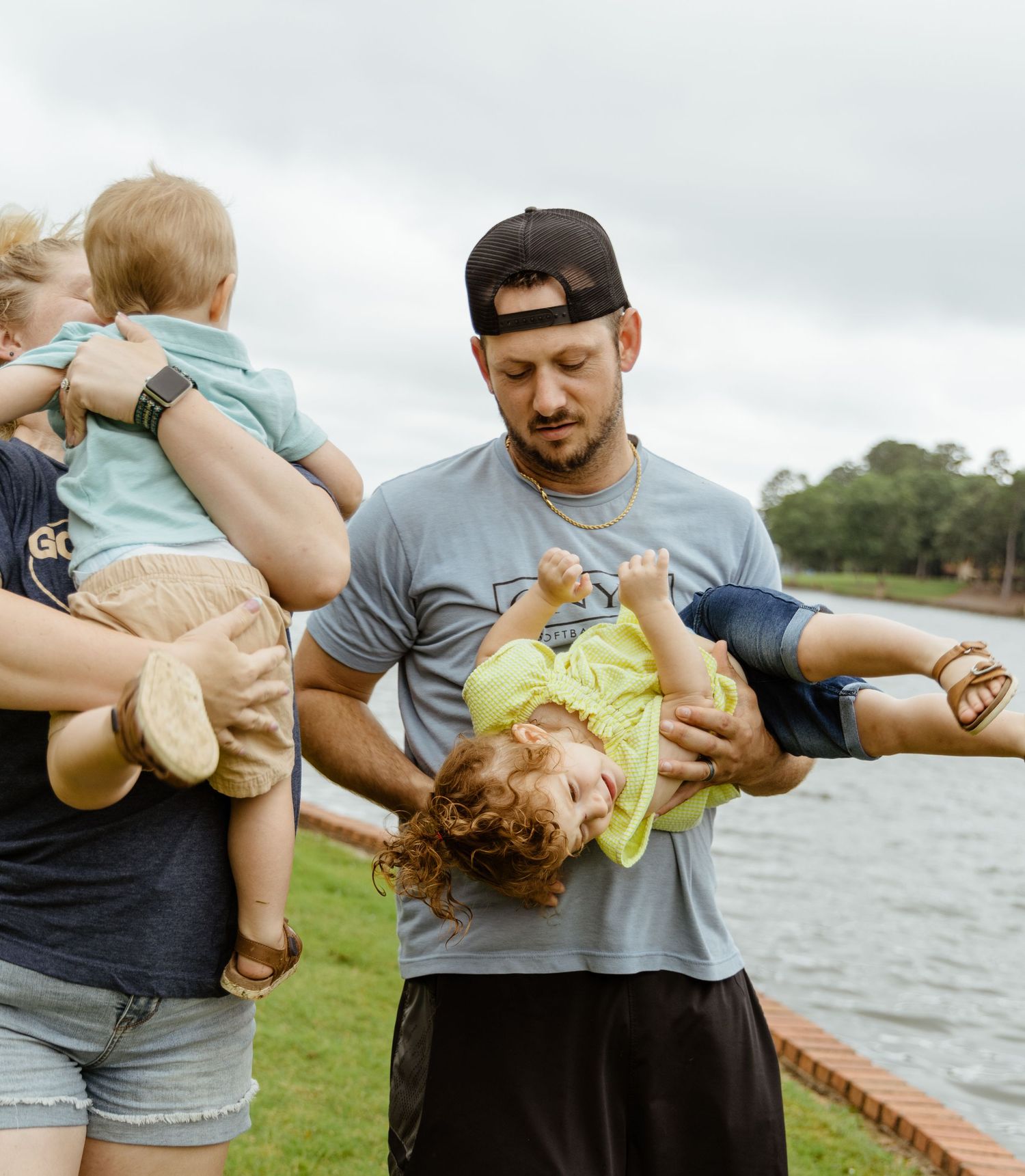 Two adults playfully lift and hold small children by a lakeside on a cloudy day.