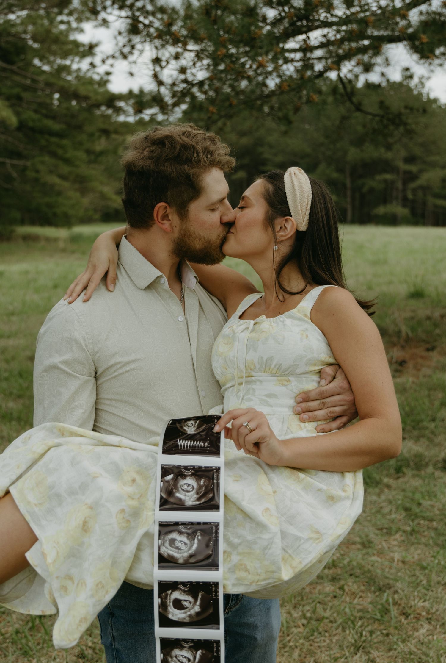 A couple shares intimate moments while holding ultrasound photos in an outdoor setting.