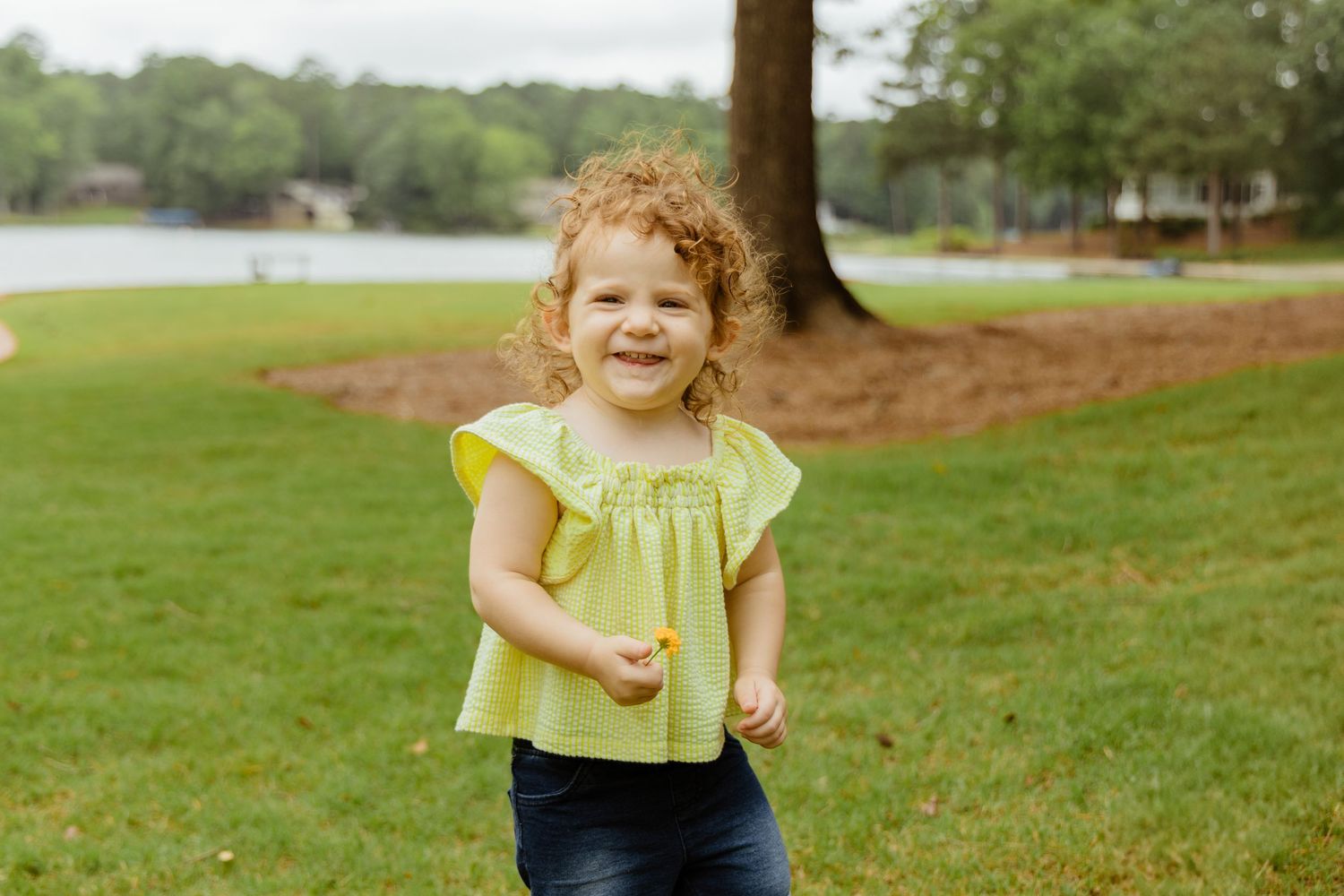 Child in yellow shirt running and playing on green grass near a lake.