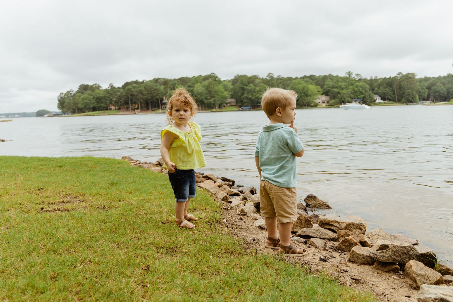 Two children play together on grassy lakeside with rocks and water in background.