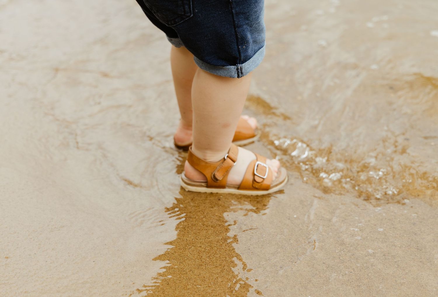 Brown sandal-clad feet splashing in shallow water on sandy shore.