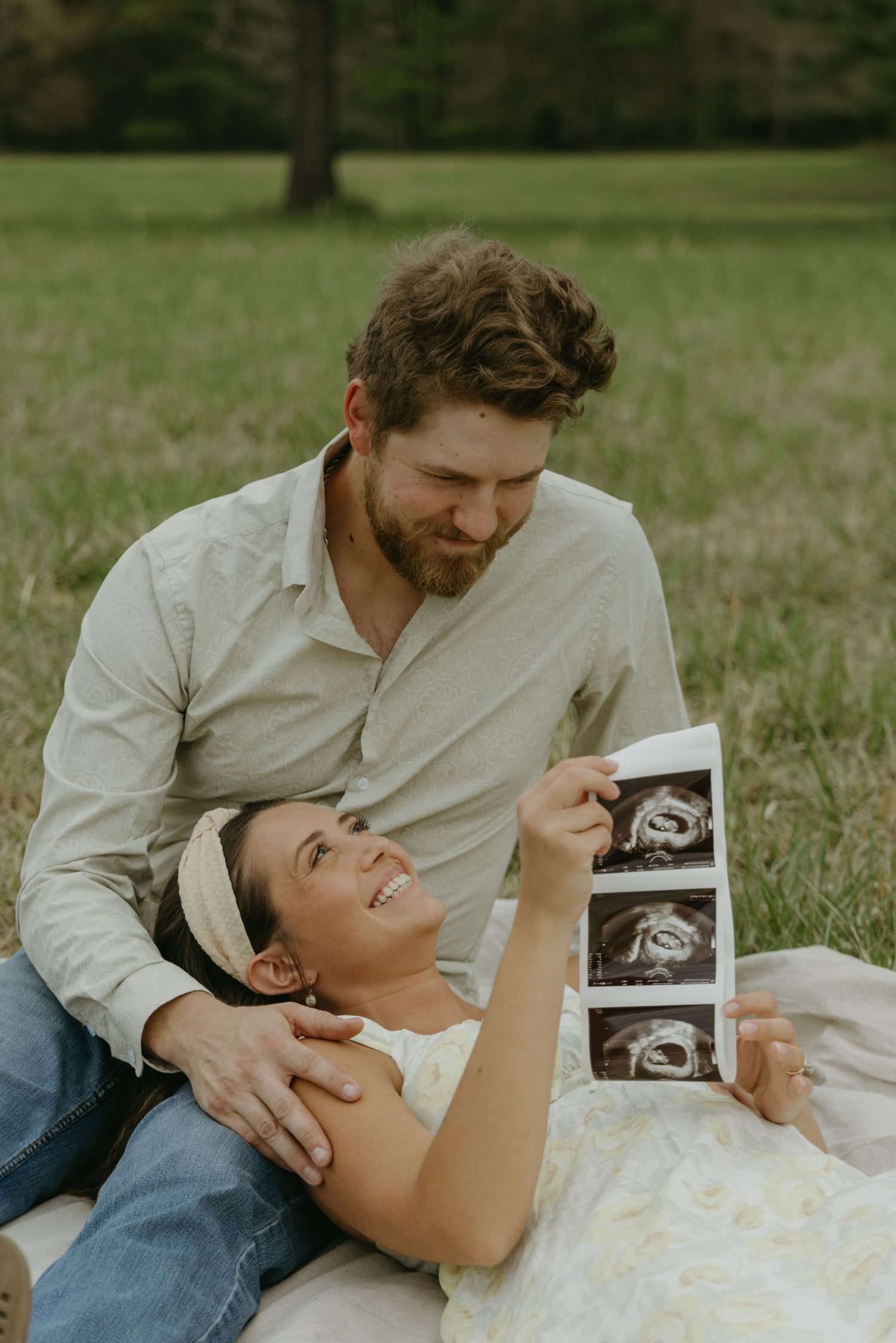 A couple lies in grass sharing joyful moments while holding ultrasound photos.