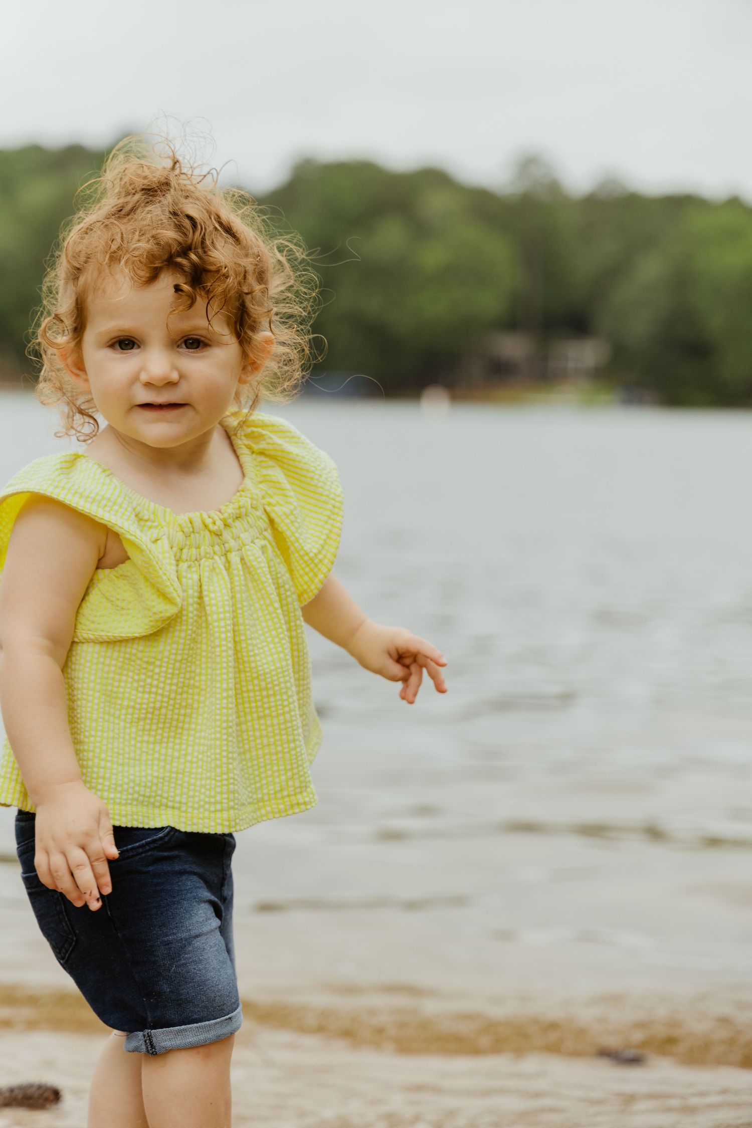 Child in yellow top plays near lakeside on sunny day.