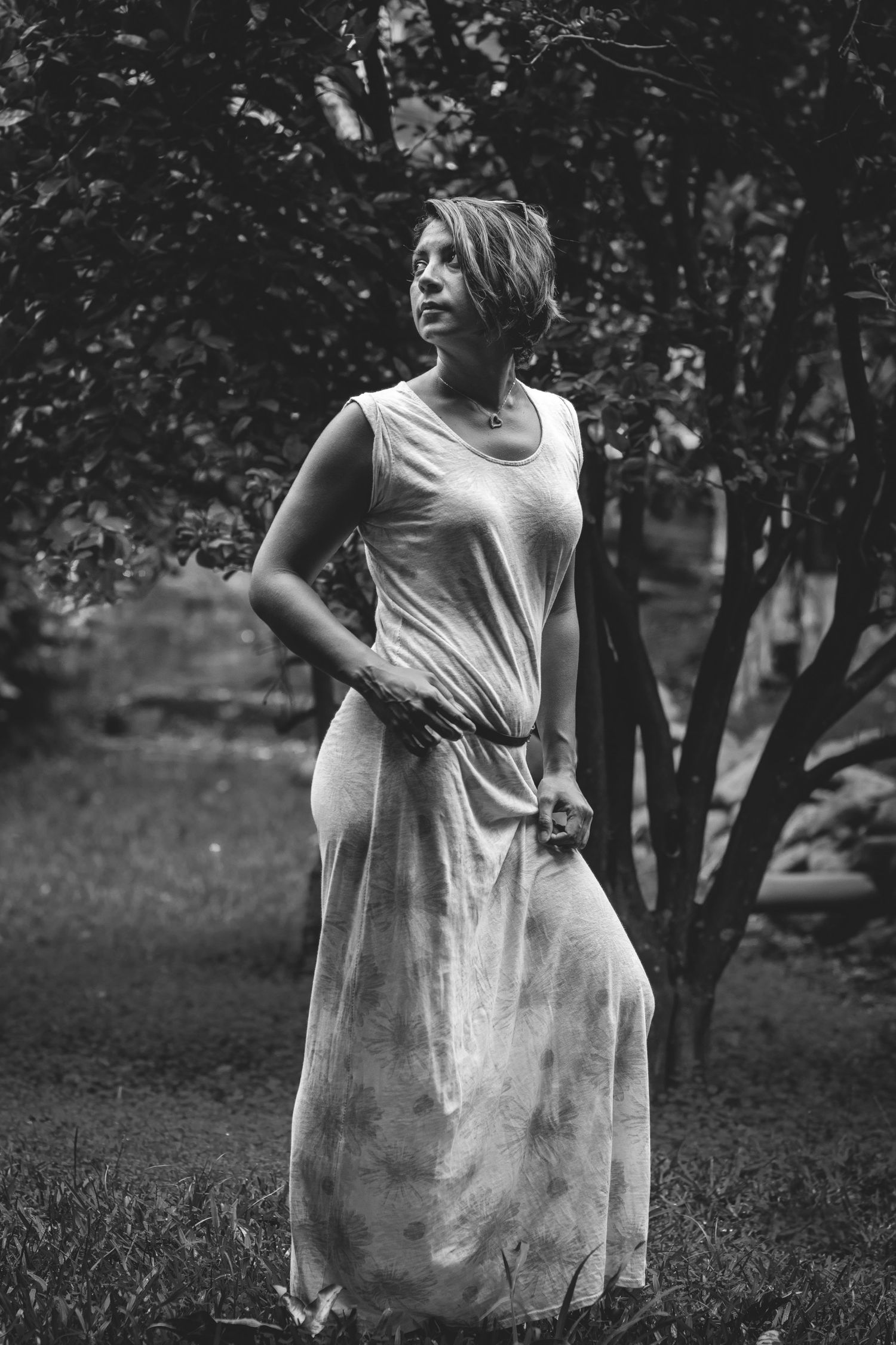A figure in flowing white dress poses thoughtfully in a garden setting captured in black and white.