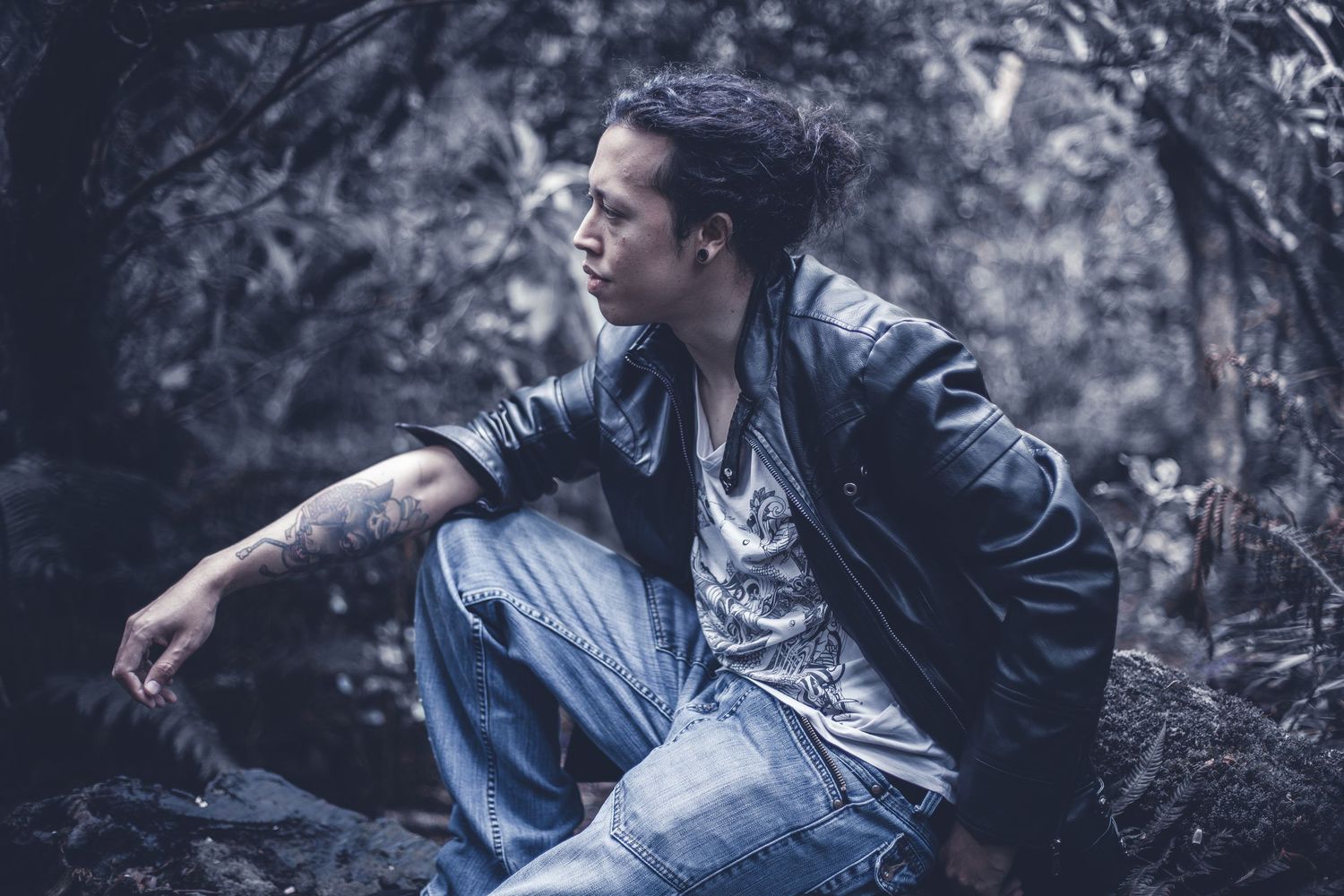 A person in leather jacket and jeans sits casually on a rock in a moody forest setting.