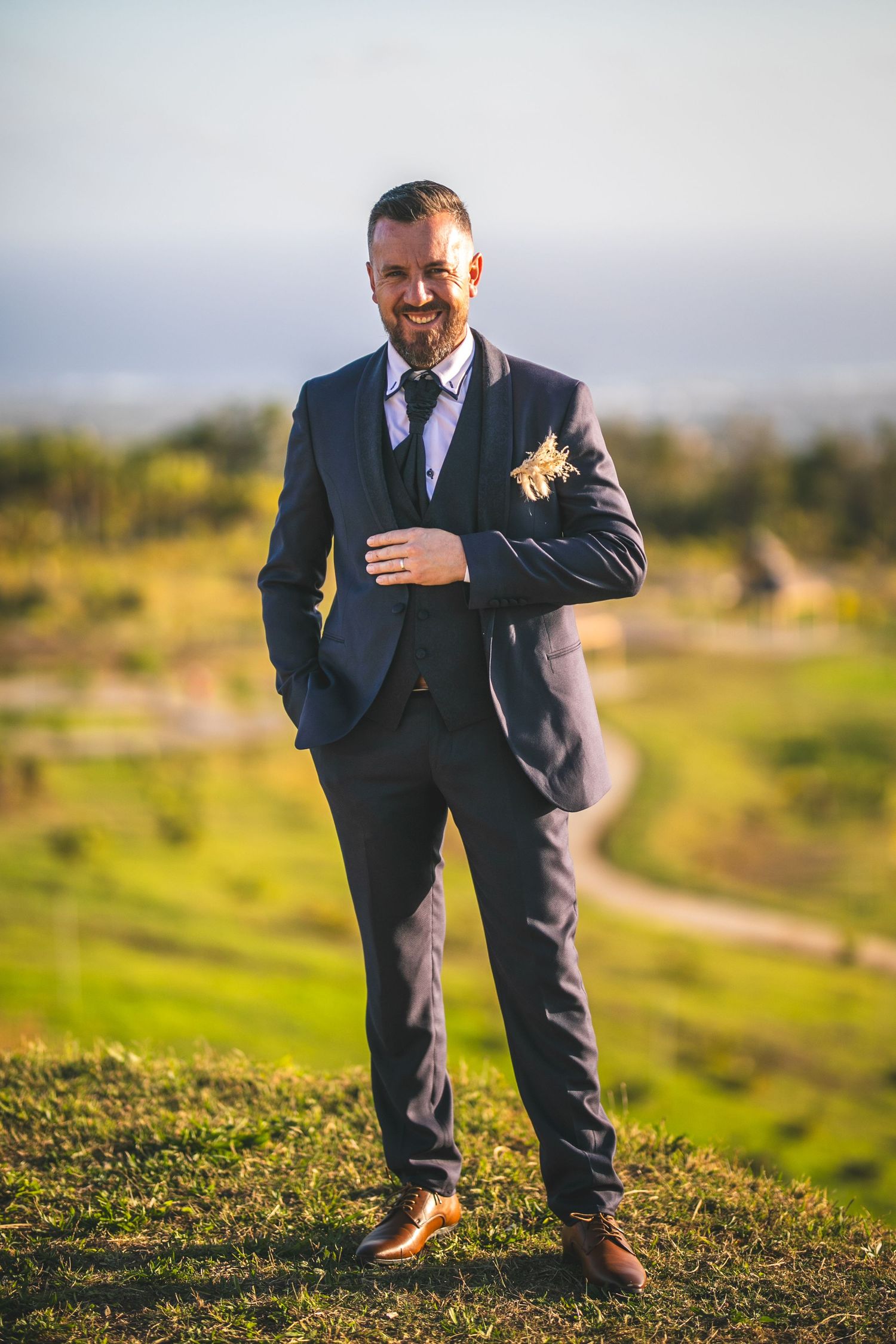 Groom in navy suit with boutonniere smiles confidently on hillside during outdoor wedding photoshoot.