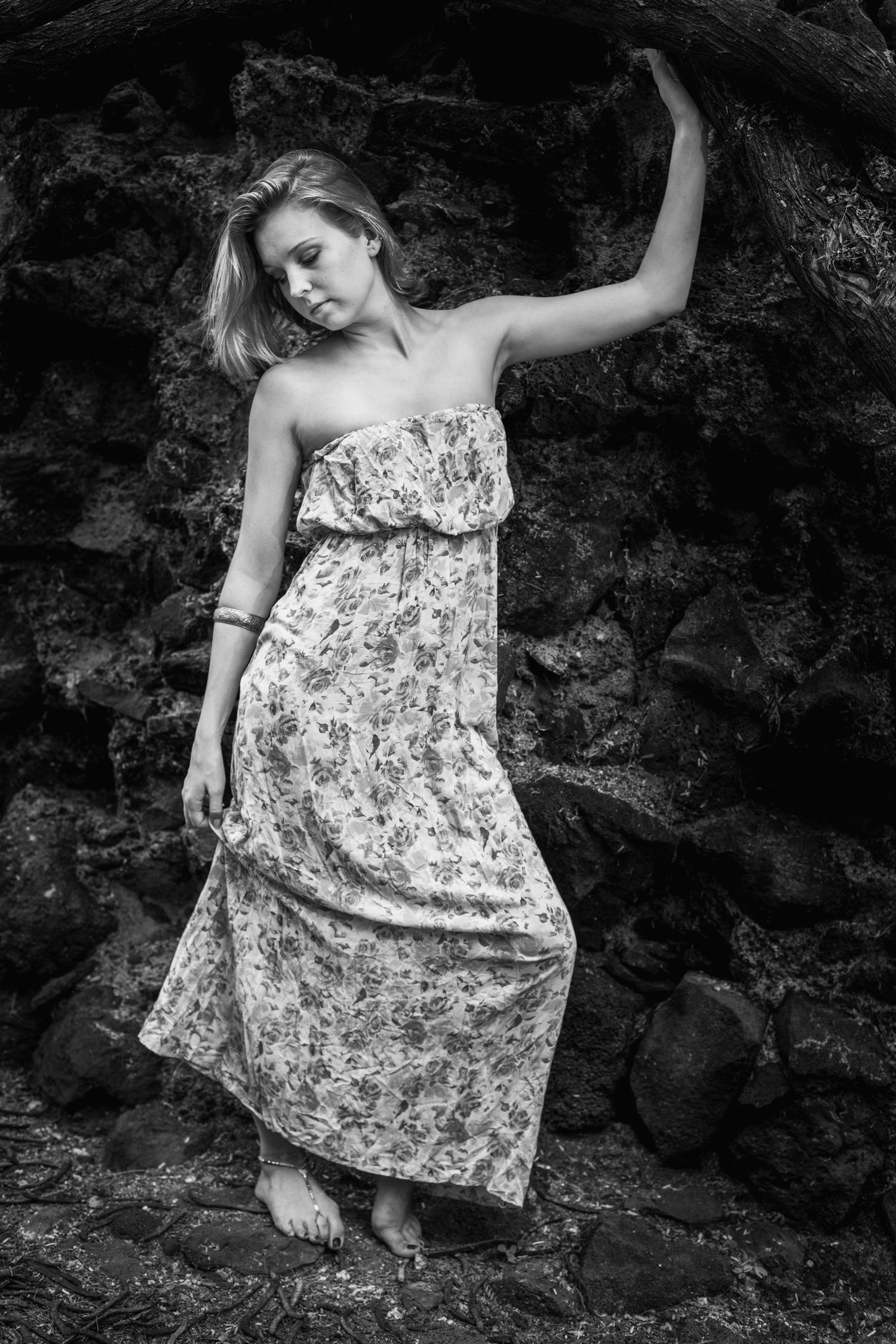 Dramatic black and white photo of model in floral maxi dress posing against rocky backdrop.