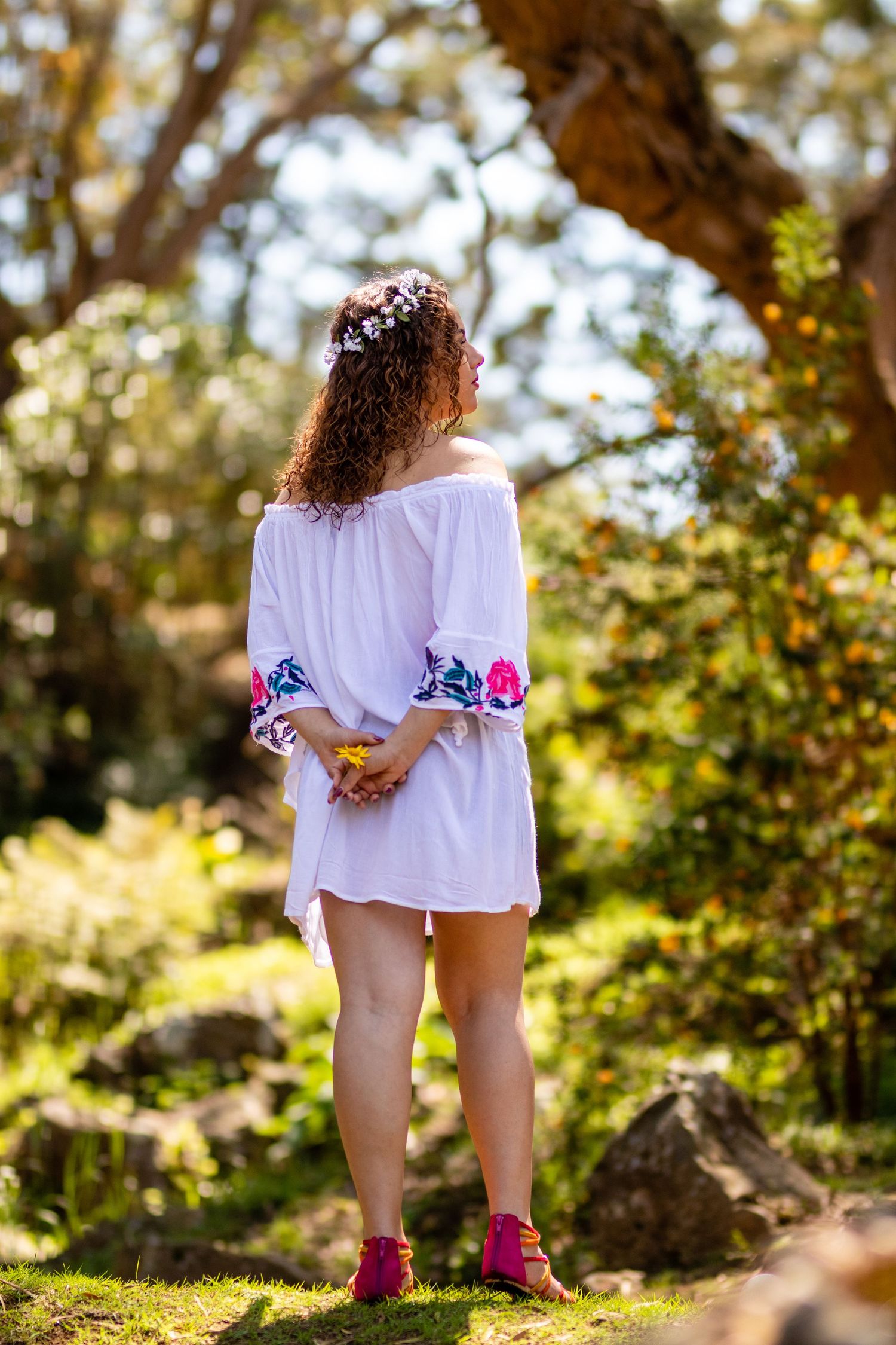 A person in white embroidered dress stands with hands behind back in sunny woodland setting.