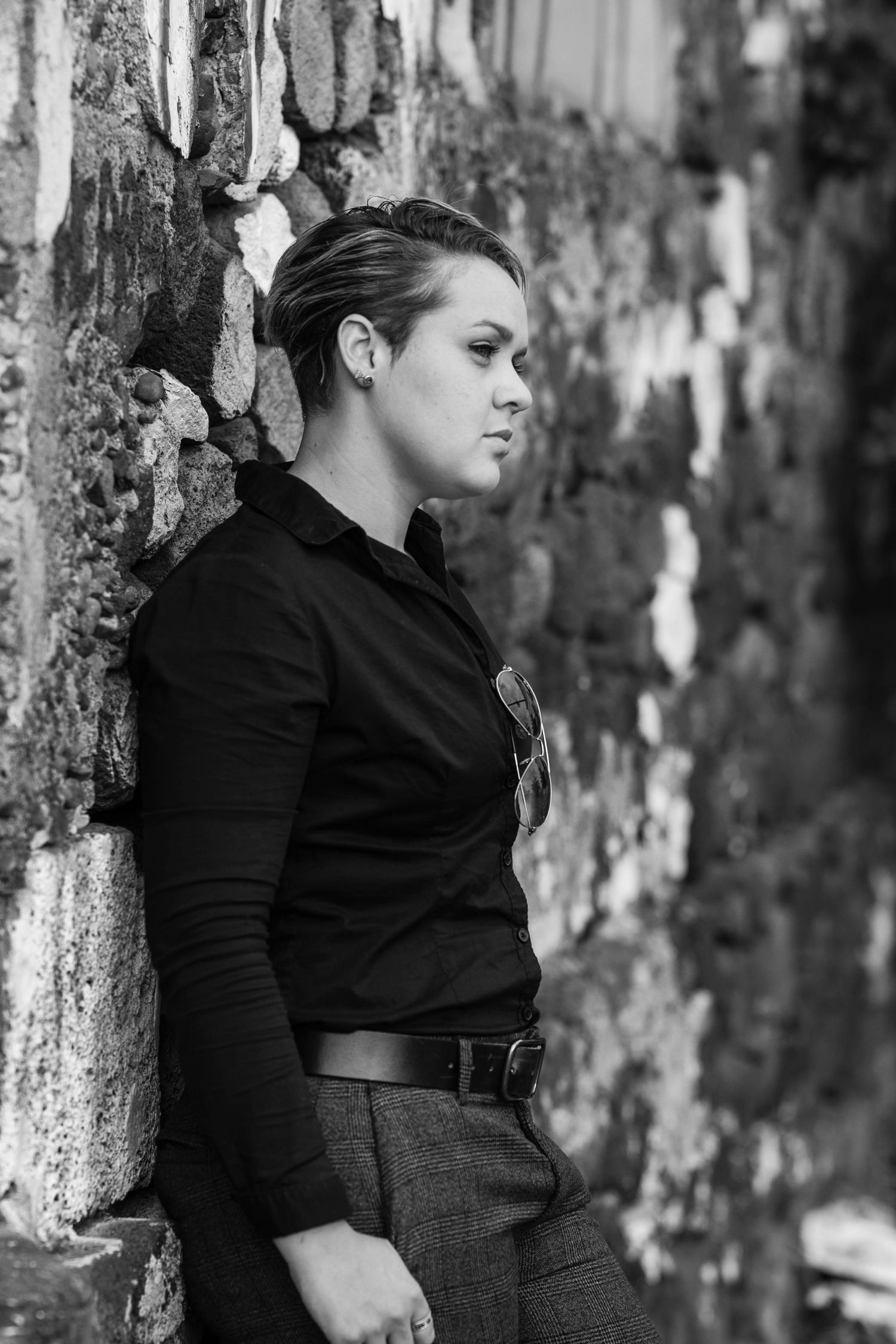 A person in black attire poses against a textured stone wall in dramatic black and white.