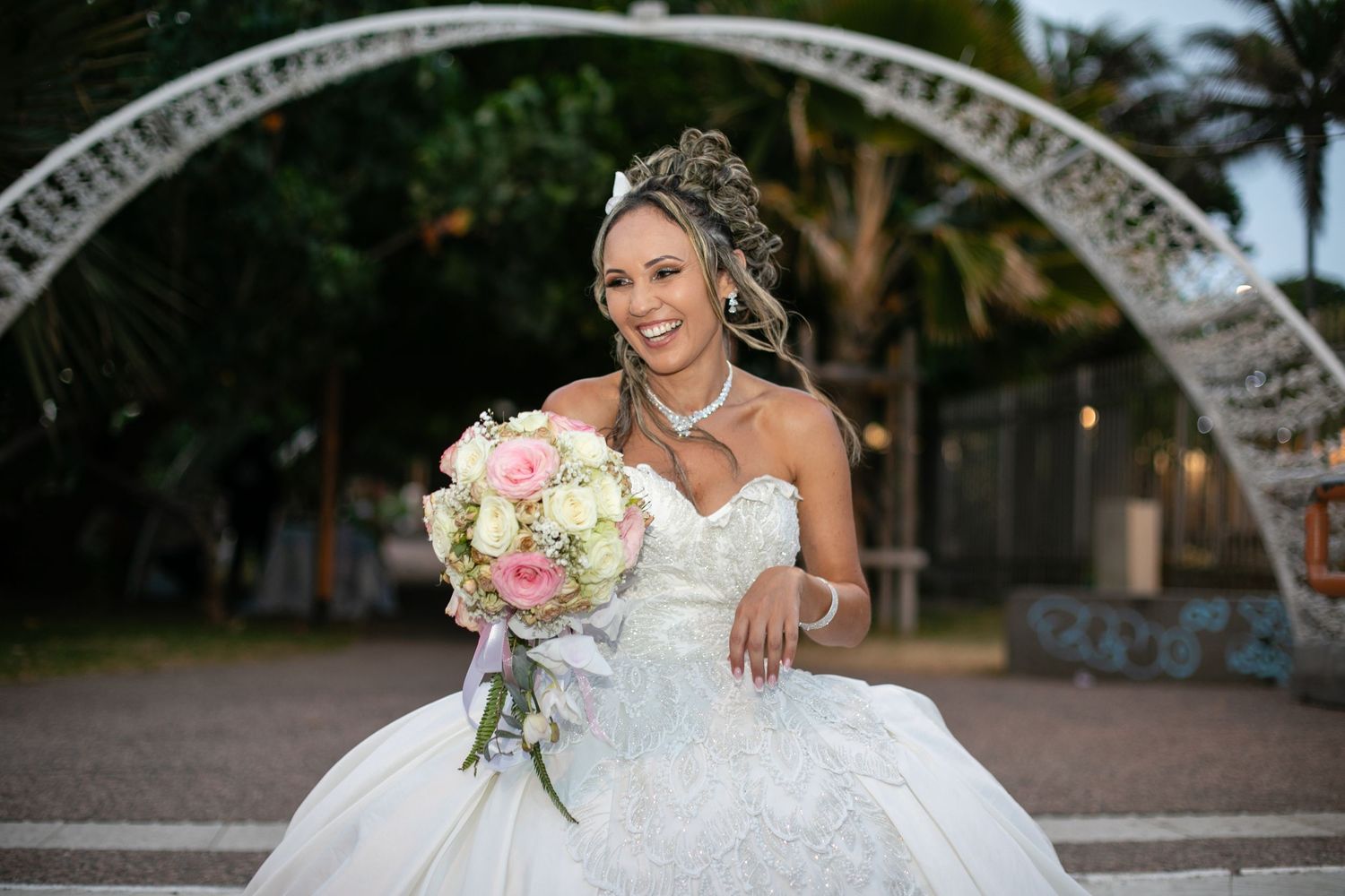 A bride in a white ball gown posing under a decorative arch with a pink bouquet.