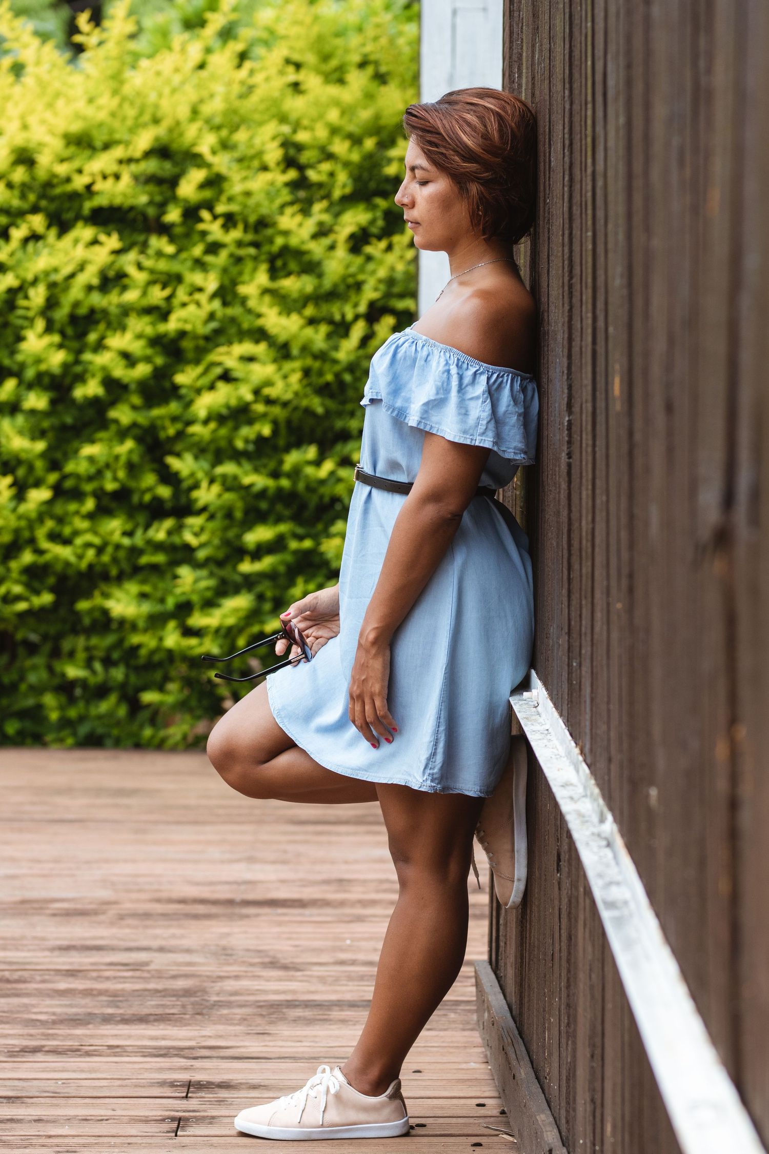 A person in light blue off-shoulder dress leans against wooden railing on outdoor deck.