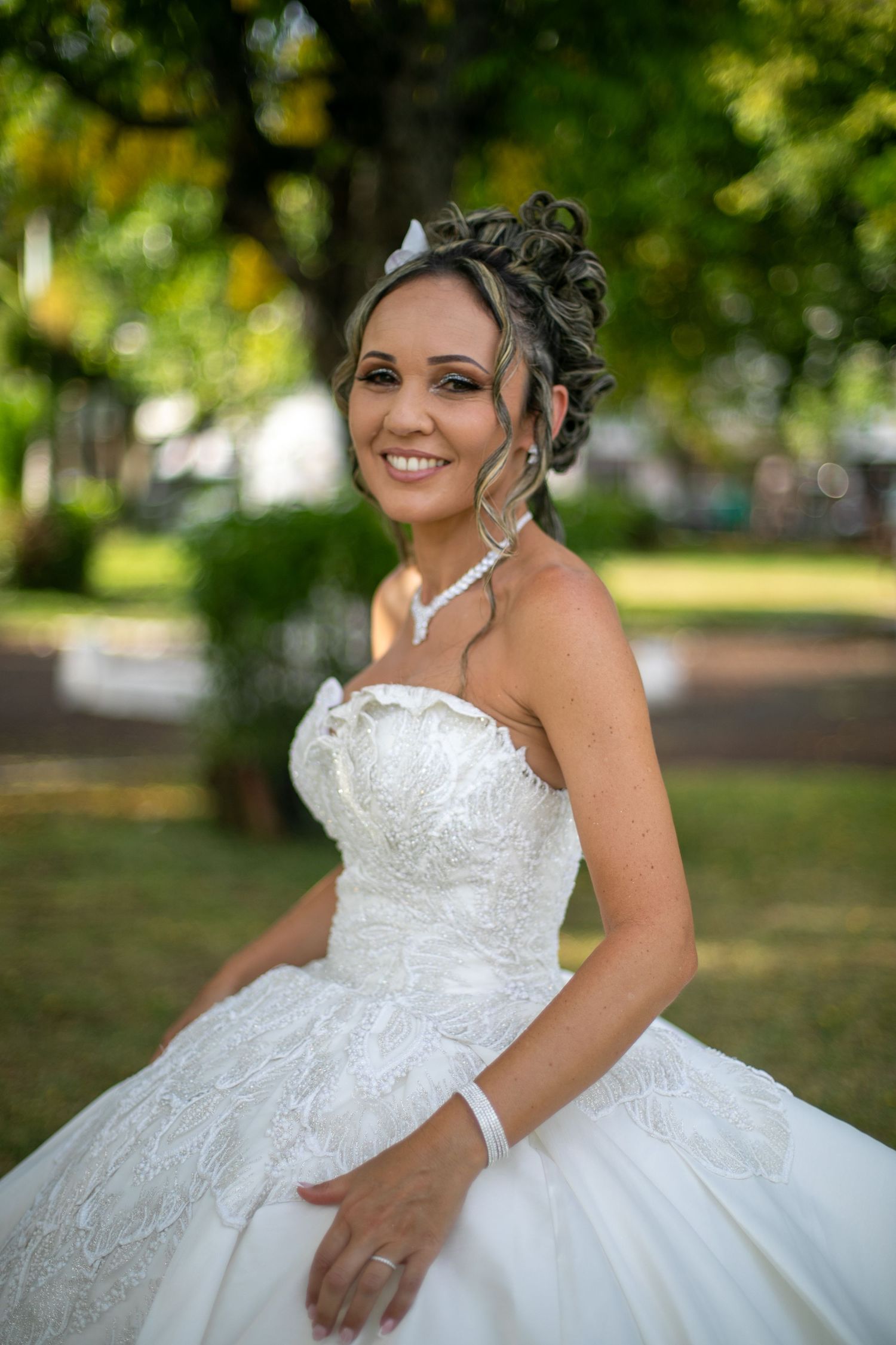 A bride in a strapless lace wedding gown smiling in a garden setting.