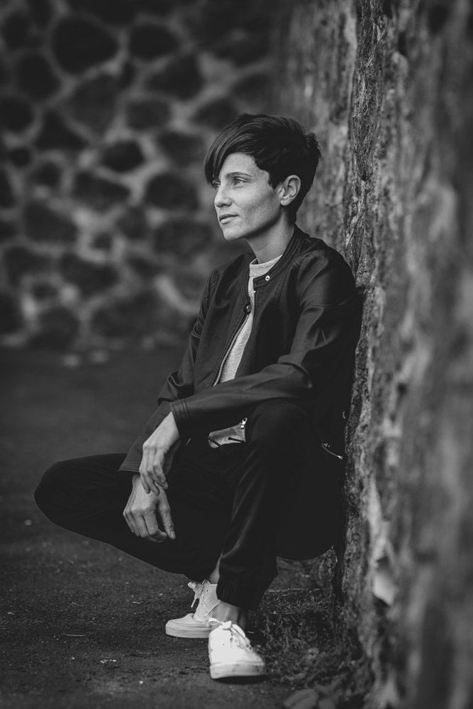 Black and white portrait of person sitting against textured stone wall in casual attire.